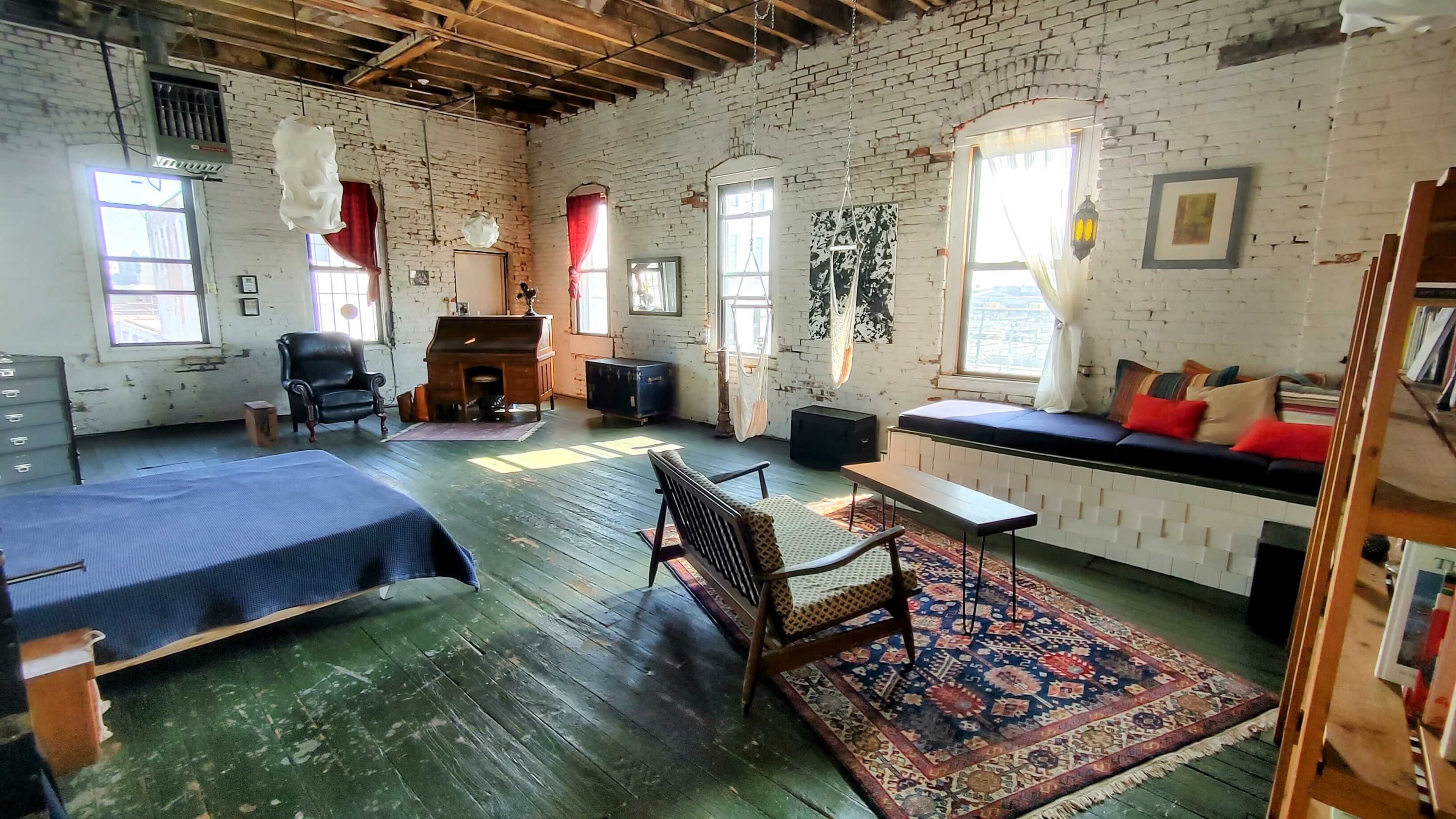 The image shows a spacious, rustic bedroom with exposed brick walls, large windows, and a mix of vintage furniture including a bed, a black chair, a wooden piano, and a patterned rug.