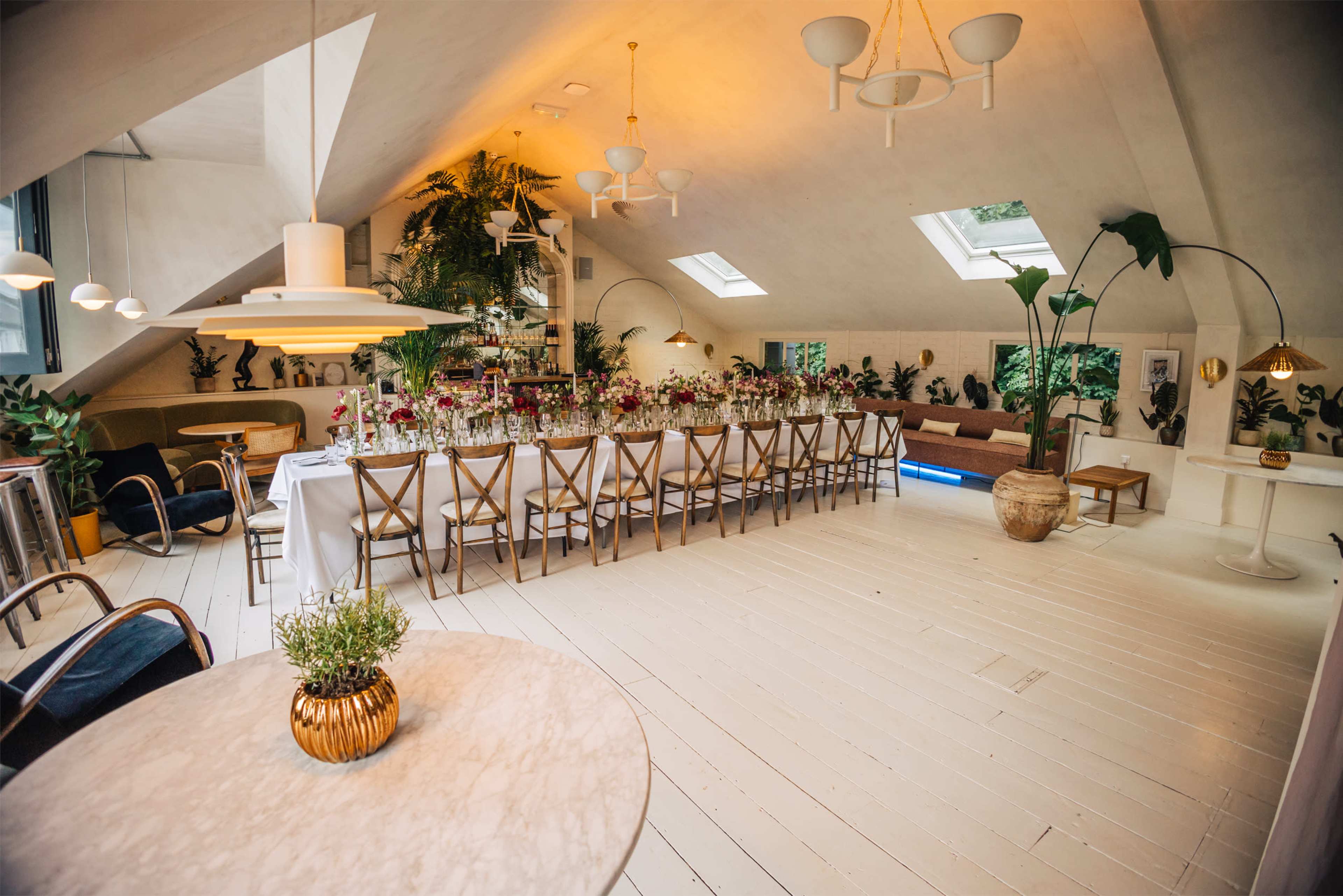 The image shows a bright, modern dining space with a long table set for an event, surrounded by plants and large windows.