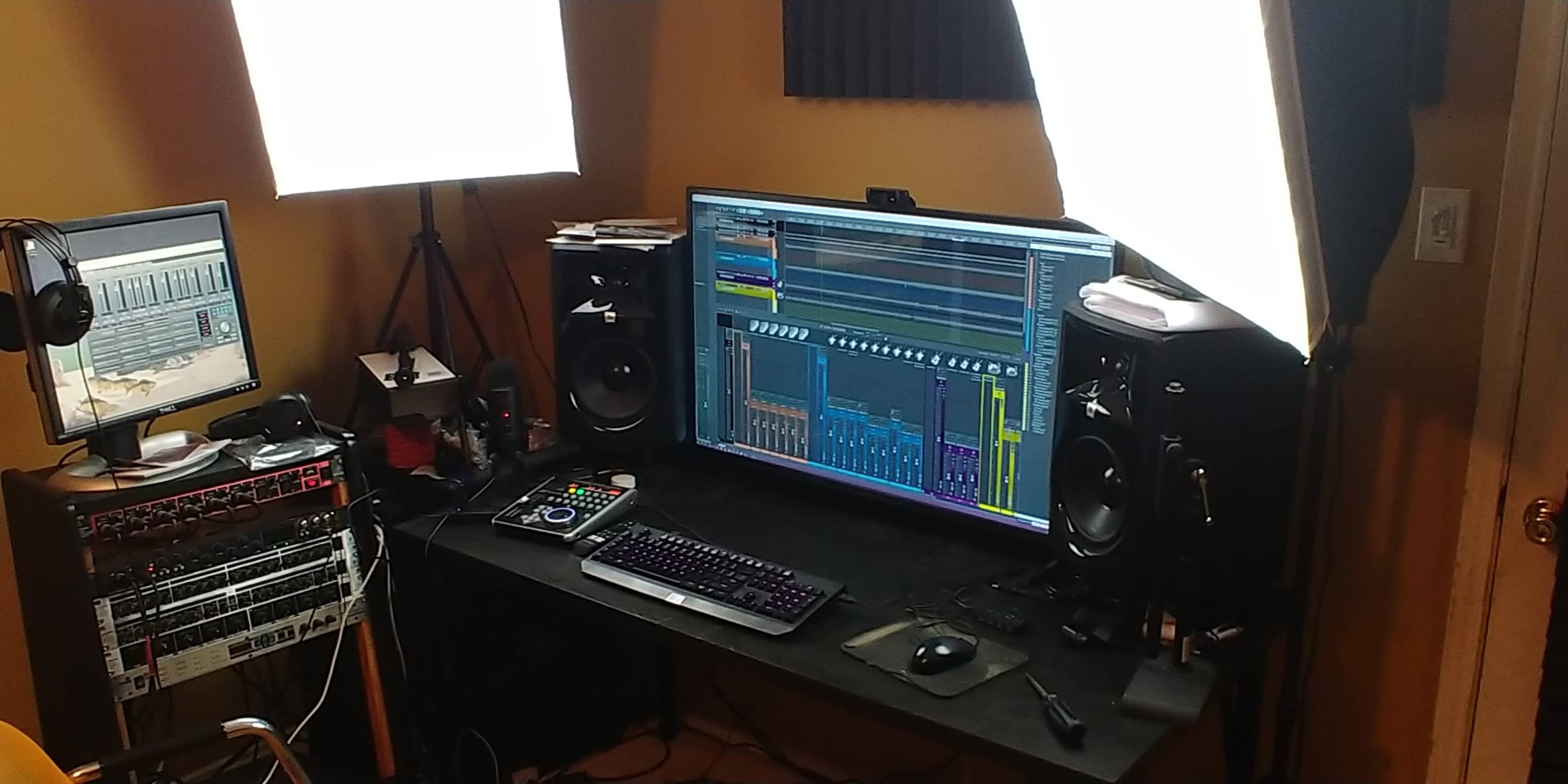 The image shows a home studio setup with a large computer monitor displaying audio software, studio monitors on either side, and a second screen beside a microphone, headphones, and recording equipment.