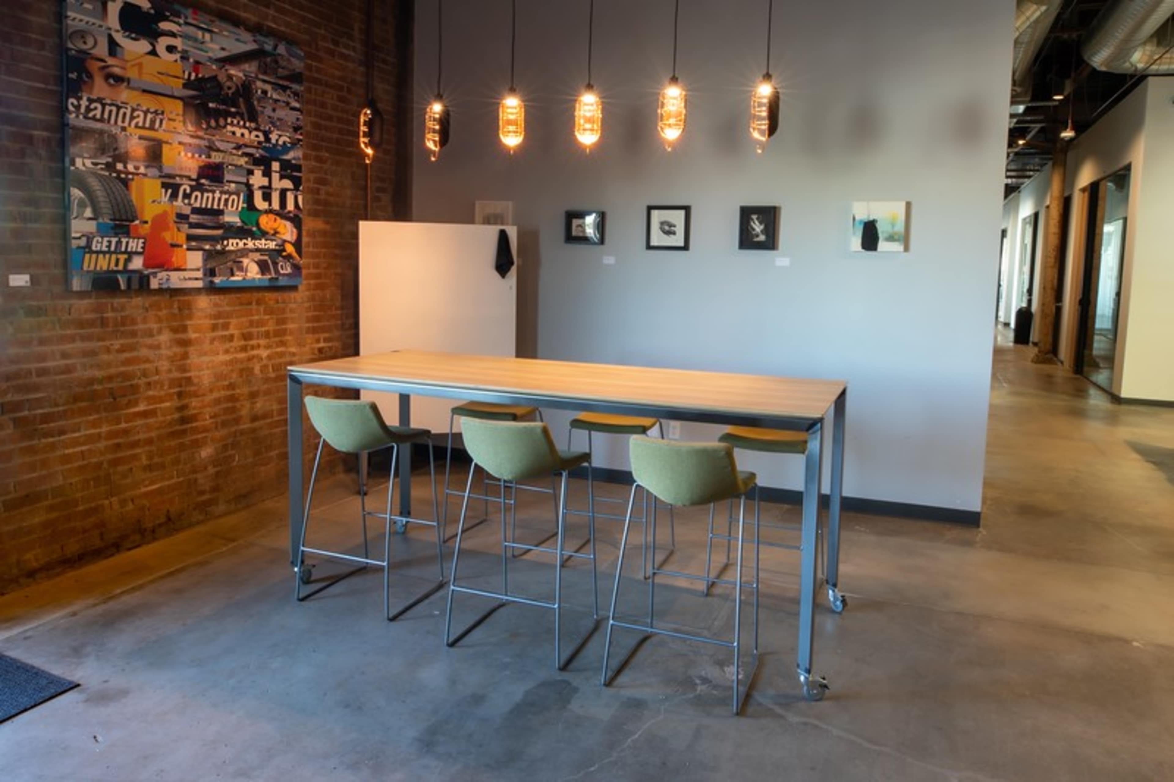A modern workspace features a wooden table with tall metal chairs, illuminated by pendant lights, against a backdrop of exposed brick and framed artwork.
