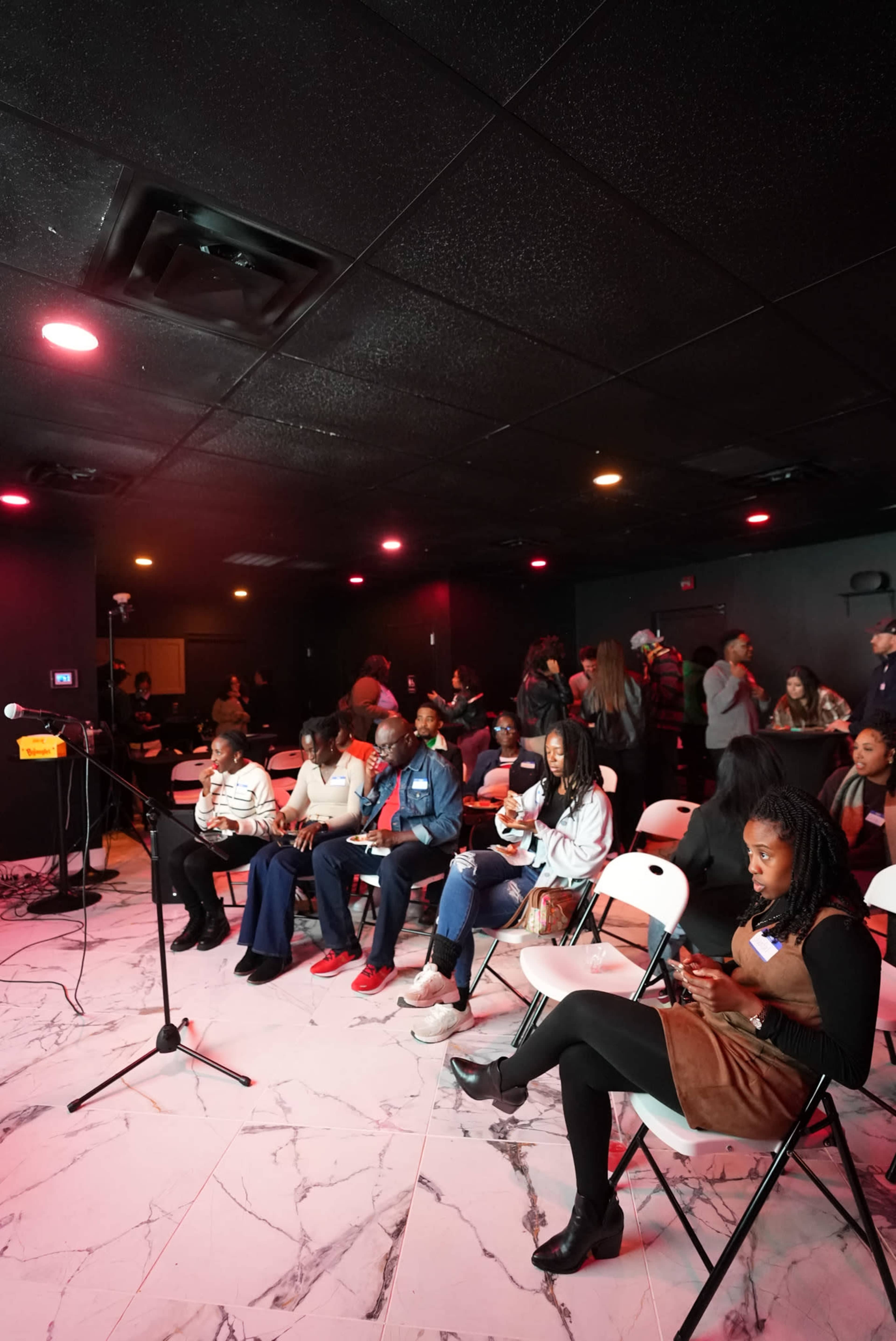 A group of people is seated in a small venue with pink lighting, some engaged with their smartphones while others mingle.