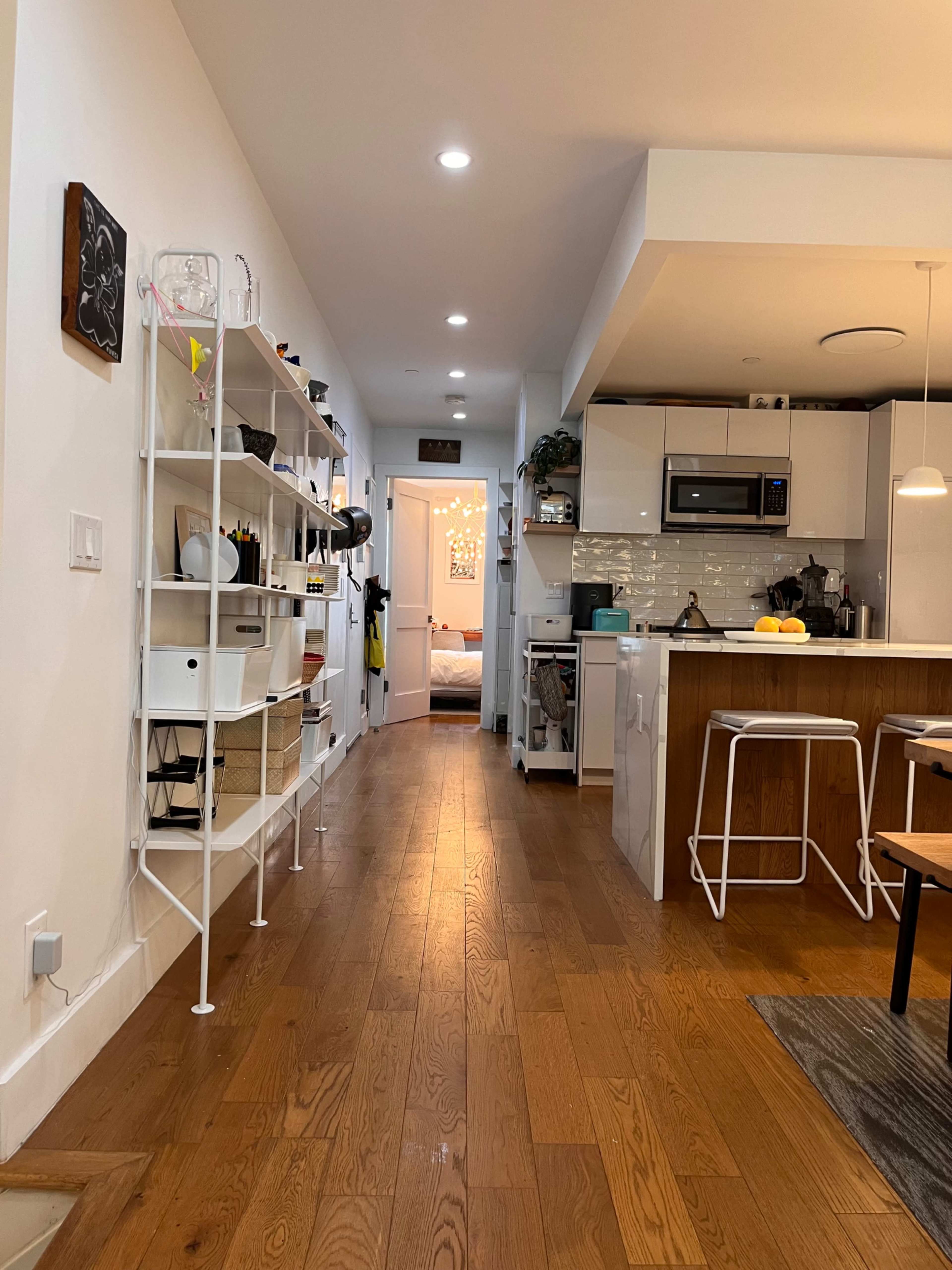 A bright, modern hallway leads to a kitchen and a door to a bedroom, with shelves on the left side displaying various items.