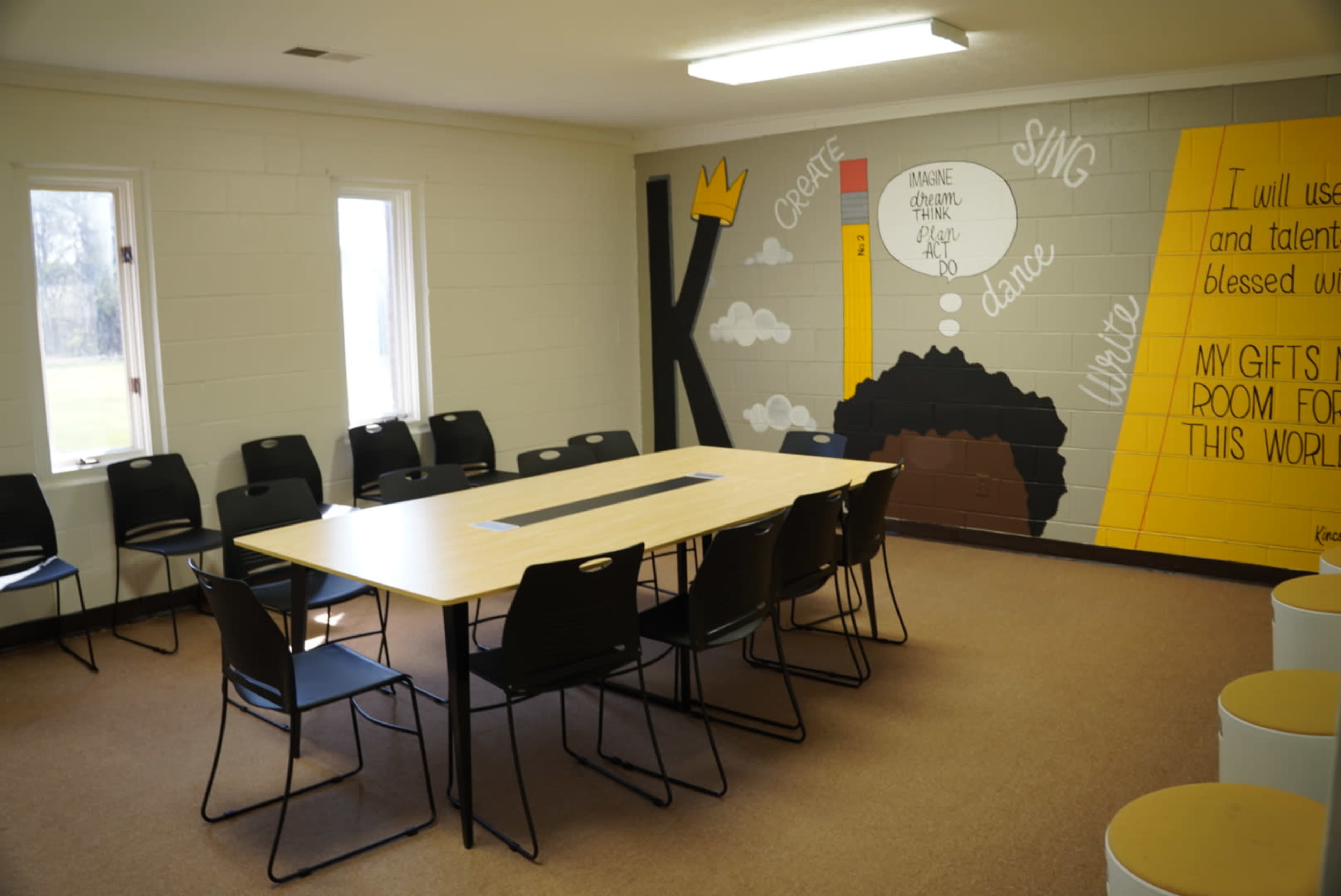 A classroom with a large table surrounded by black chairs and a colorful mural on the wall featuring motivational phrases.