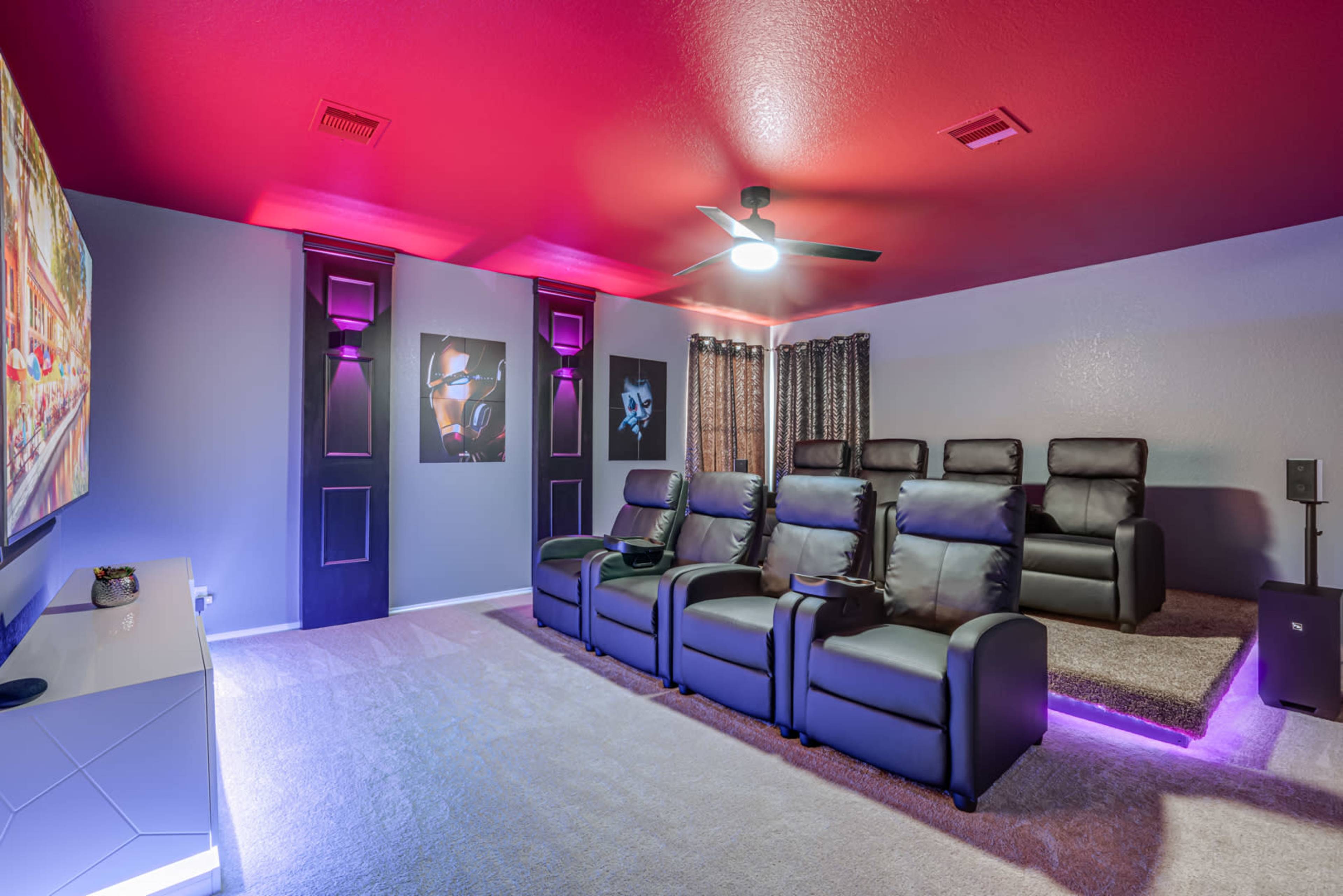 A home theater room with six black reclining chairs, a large TV mounted on the wall, and dim lighting accentuated by purple and white features.