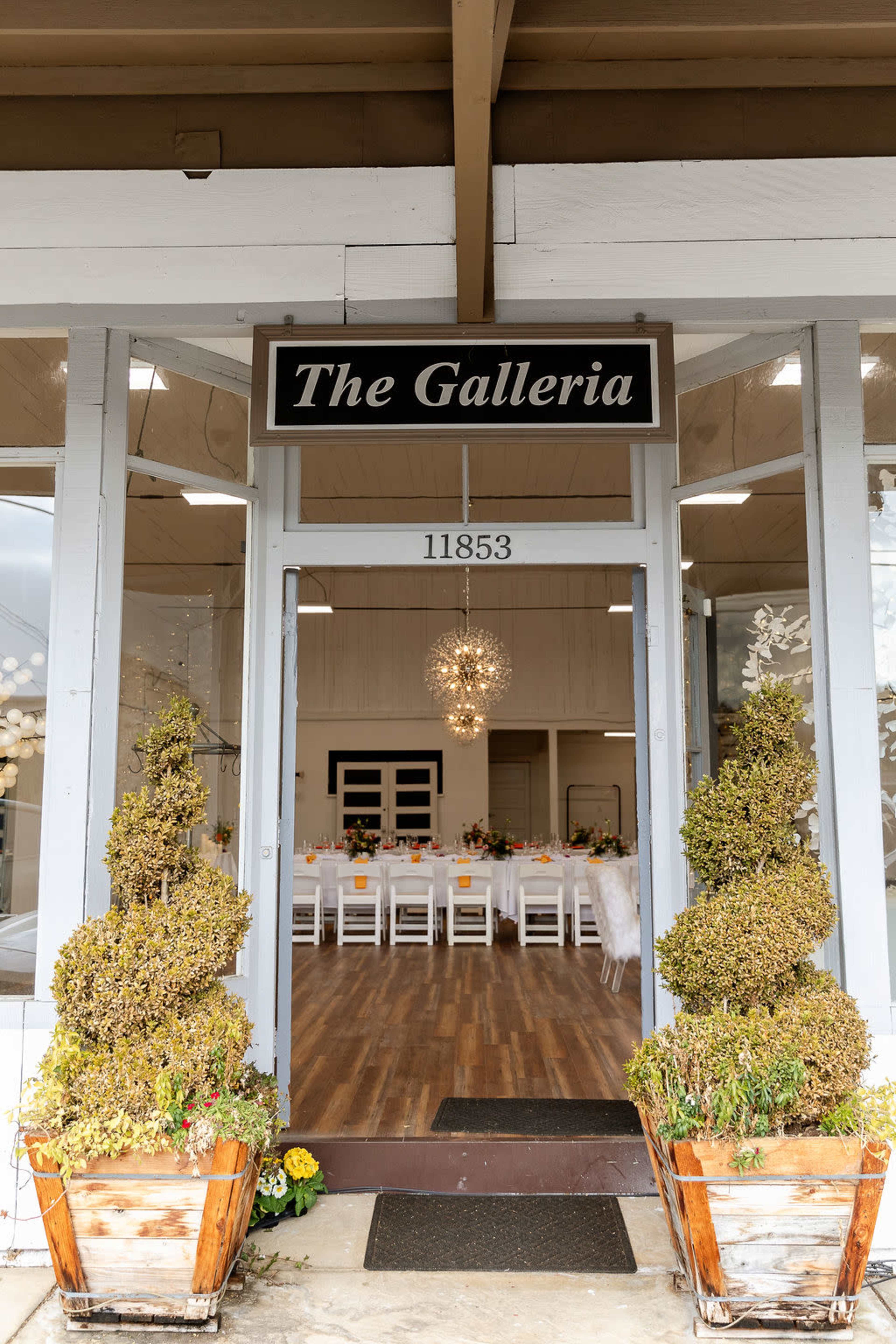 The image shows the entrance to a venue called "The Galleria," featuring two topiary shrubs in planters flanking the door, which opens to a well-lit room with a long dining table set for an event.