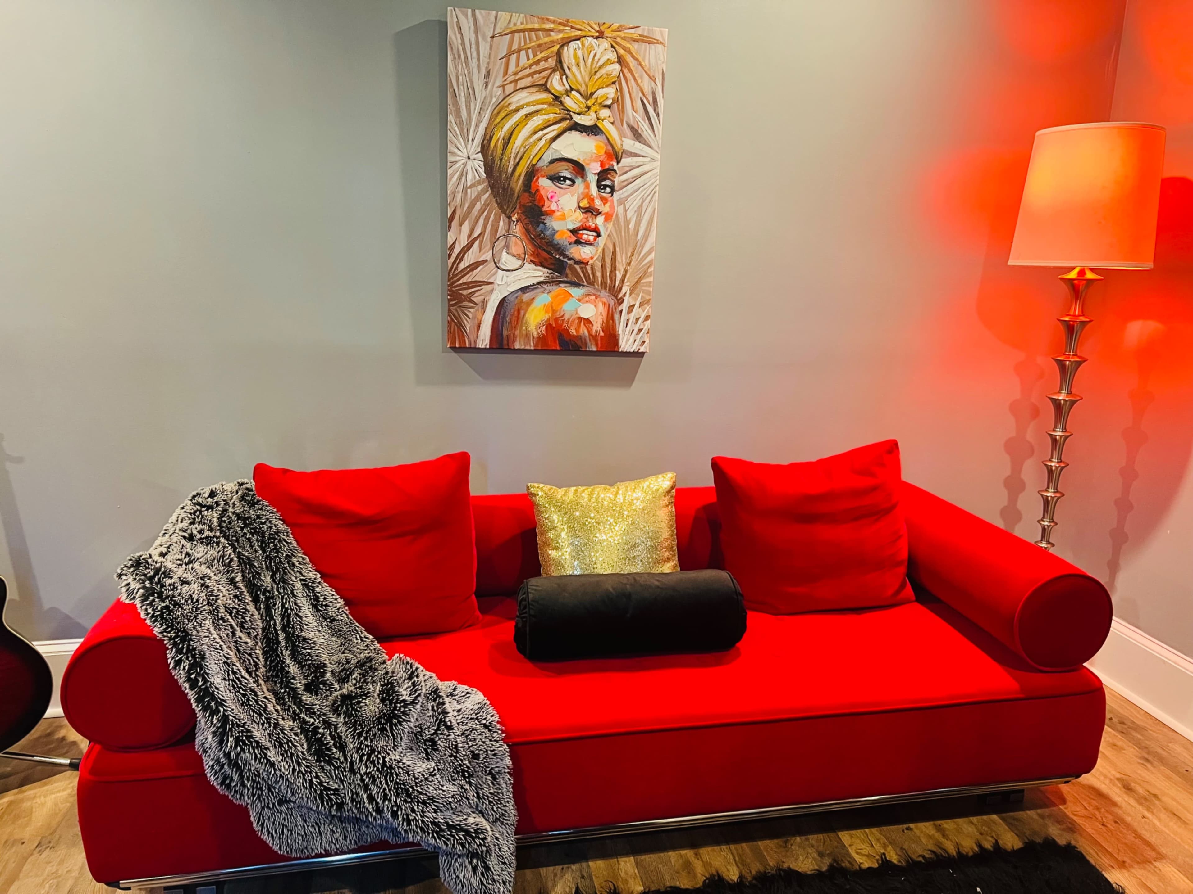 The image features a red couch with decorative pillows, a throw blanket, and a wall art piece depicting a portrait of a woman.