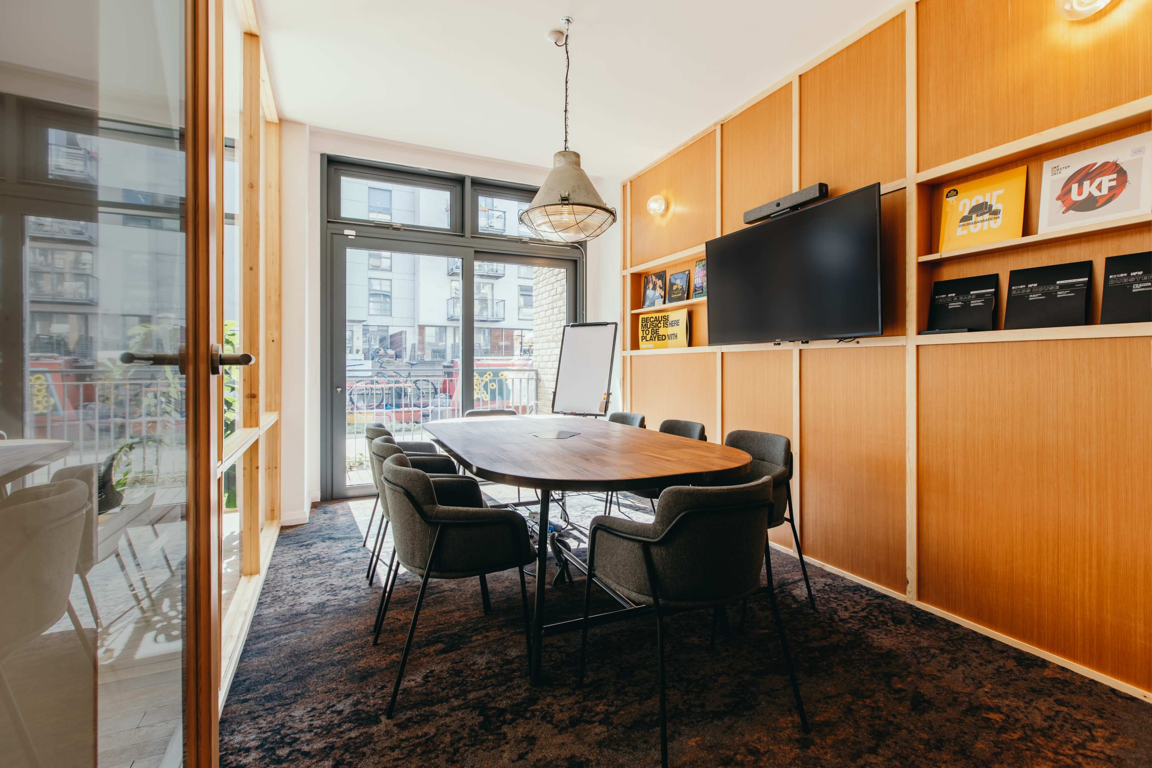 A modern conference room featuring a large wooden table surrounded by six chairs, a wall-mounted television, and shelves displaying various items.