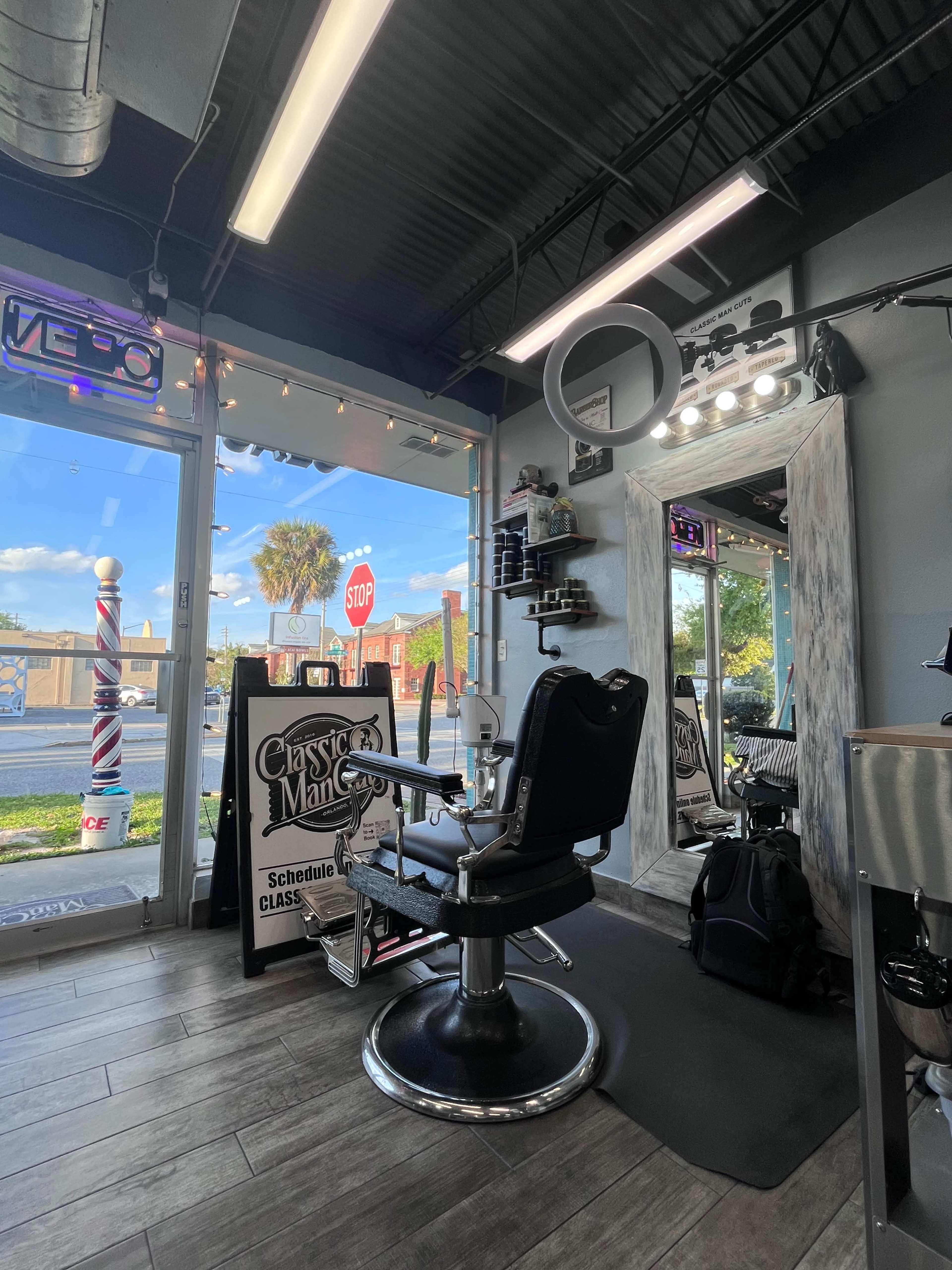 The interior of a barber shop features a black barber chair, a large mirror, and various grooming products arranged on shelves, with a view of a street outside through large windows.