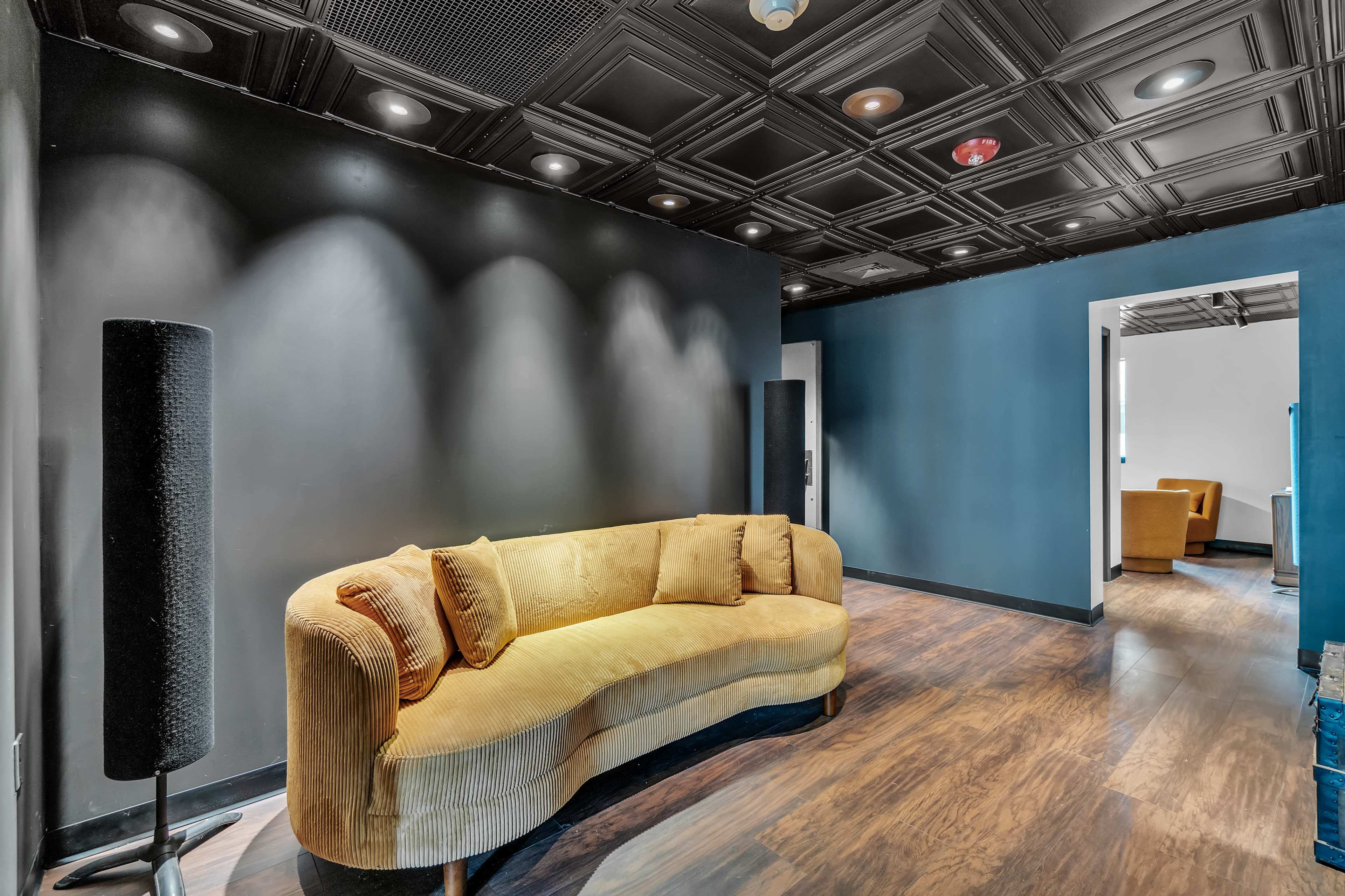 The image shows a modern lounge area with a curved yellow sofa, dark walls, and wooden flooring, alongside a doorway leading to another room.