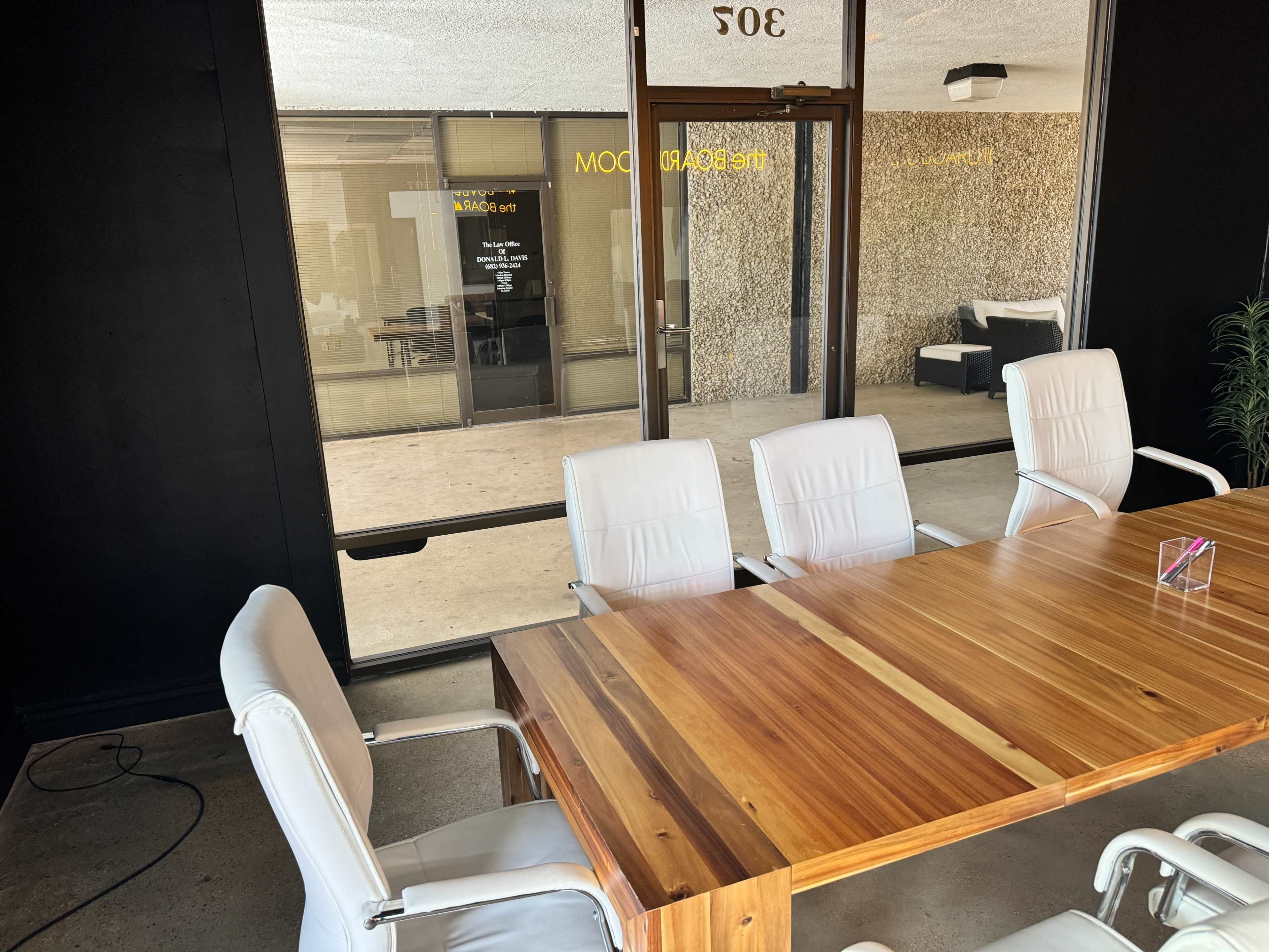 A wooden conference table with white chairs is positioned in a meeting room, facing large glass doors that lead outside.