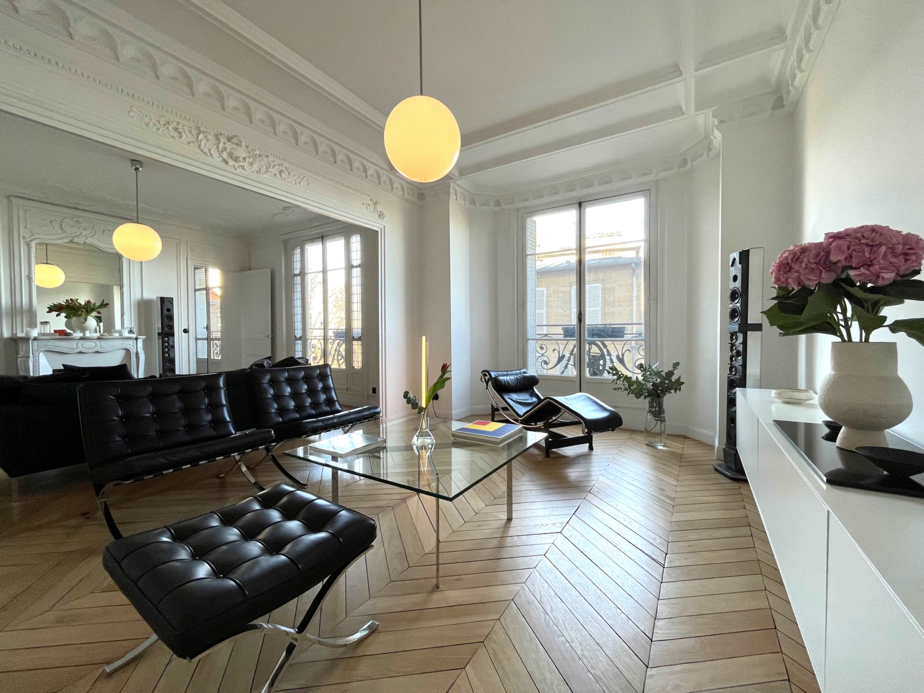 The image shows a modern living room with a glass coffee table, black lounge chairs, and a vase of flowers, featuring large windows and herringbone wooden flooring.