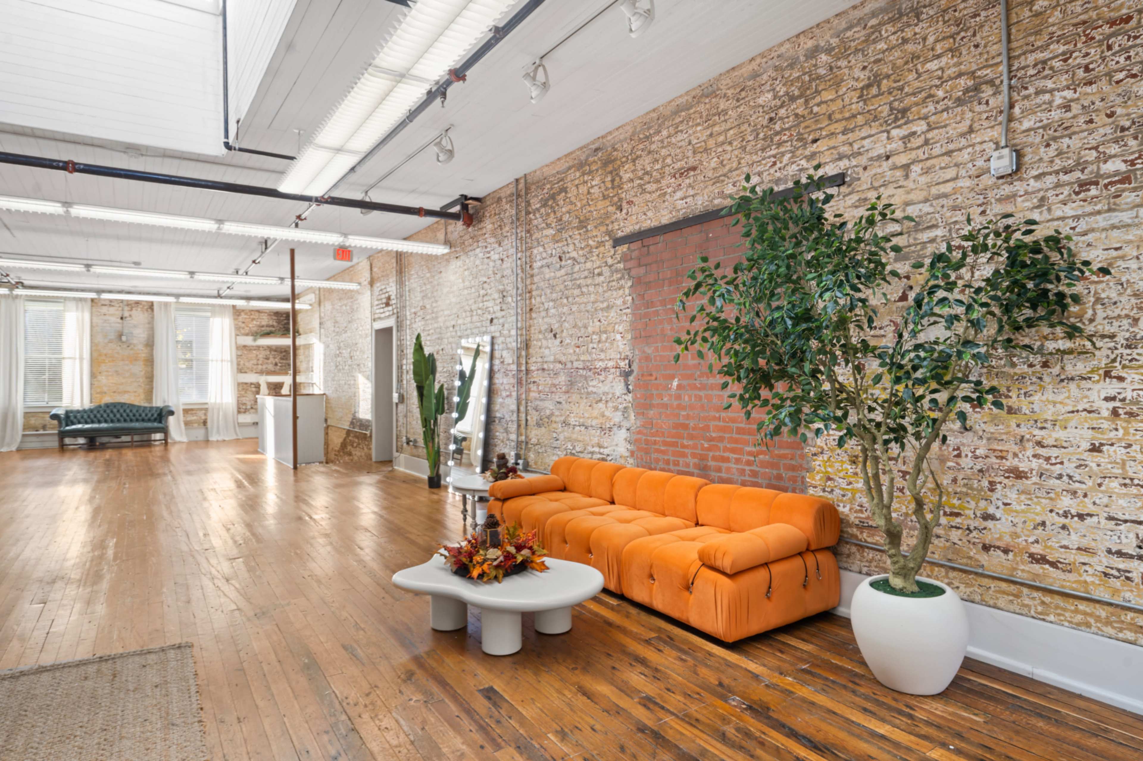 The image shows a spacious room with hardwood floors, a bright orange sofa, a potted plant, and exposed brick walls.