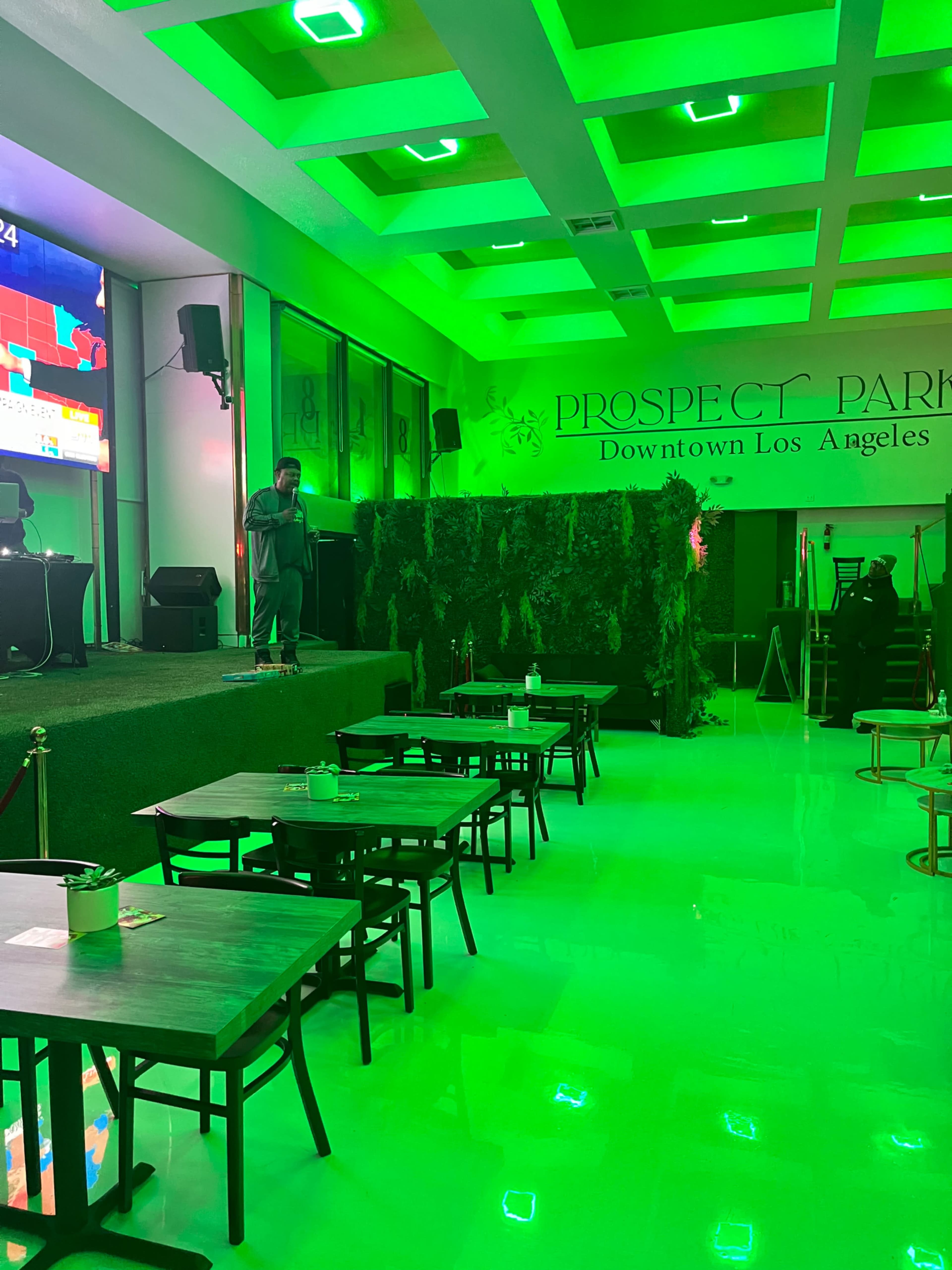 The image shows a modern indoor venue with green lighting, featuring empty tables and a stage area on one side.