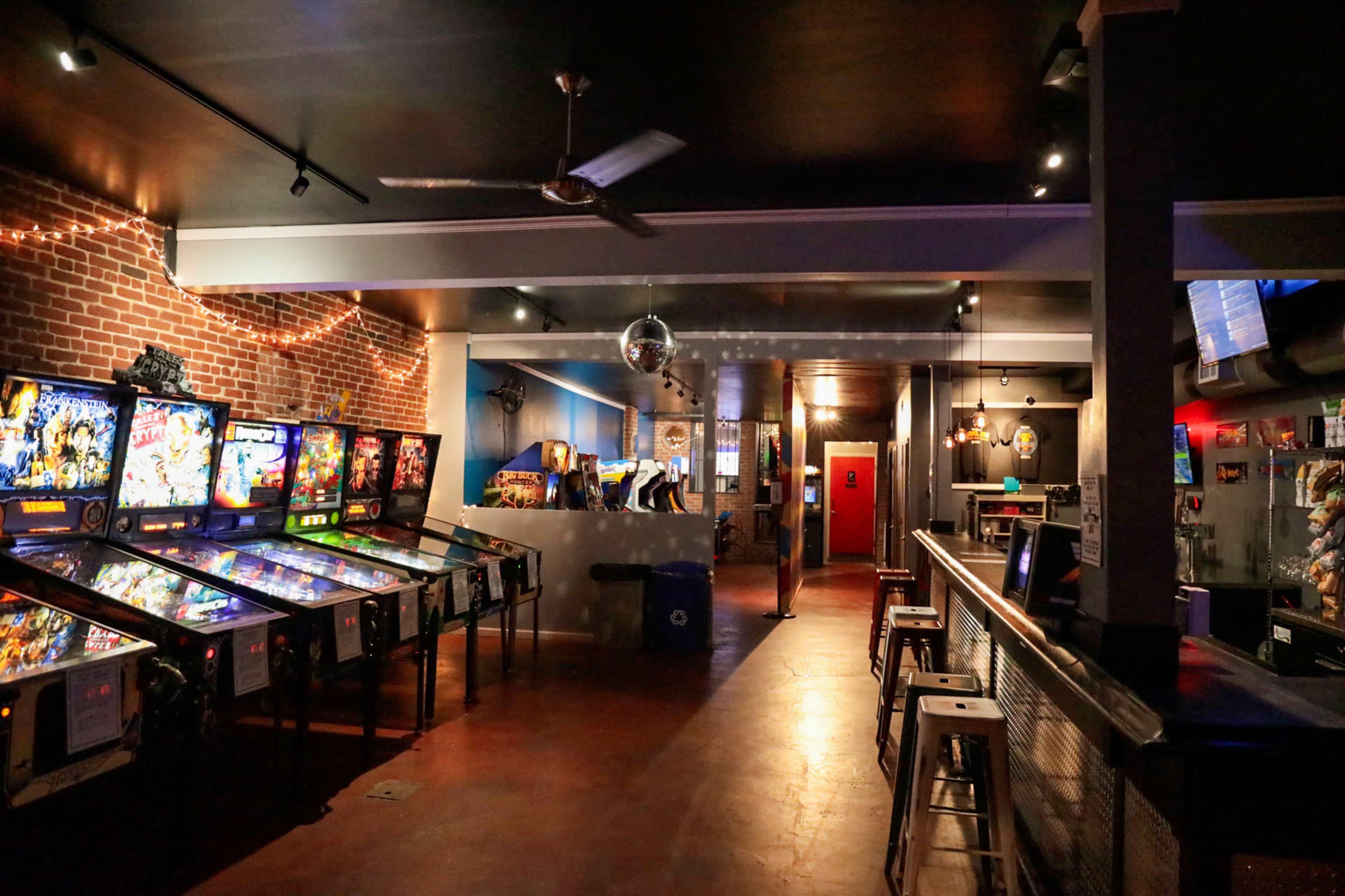 A retro arcade with pinball machines lined up against a brick wall and a bar in the background.