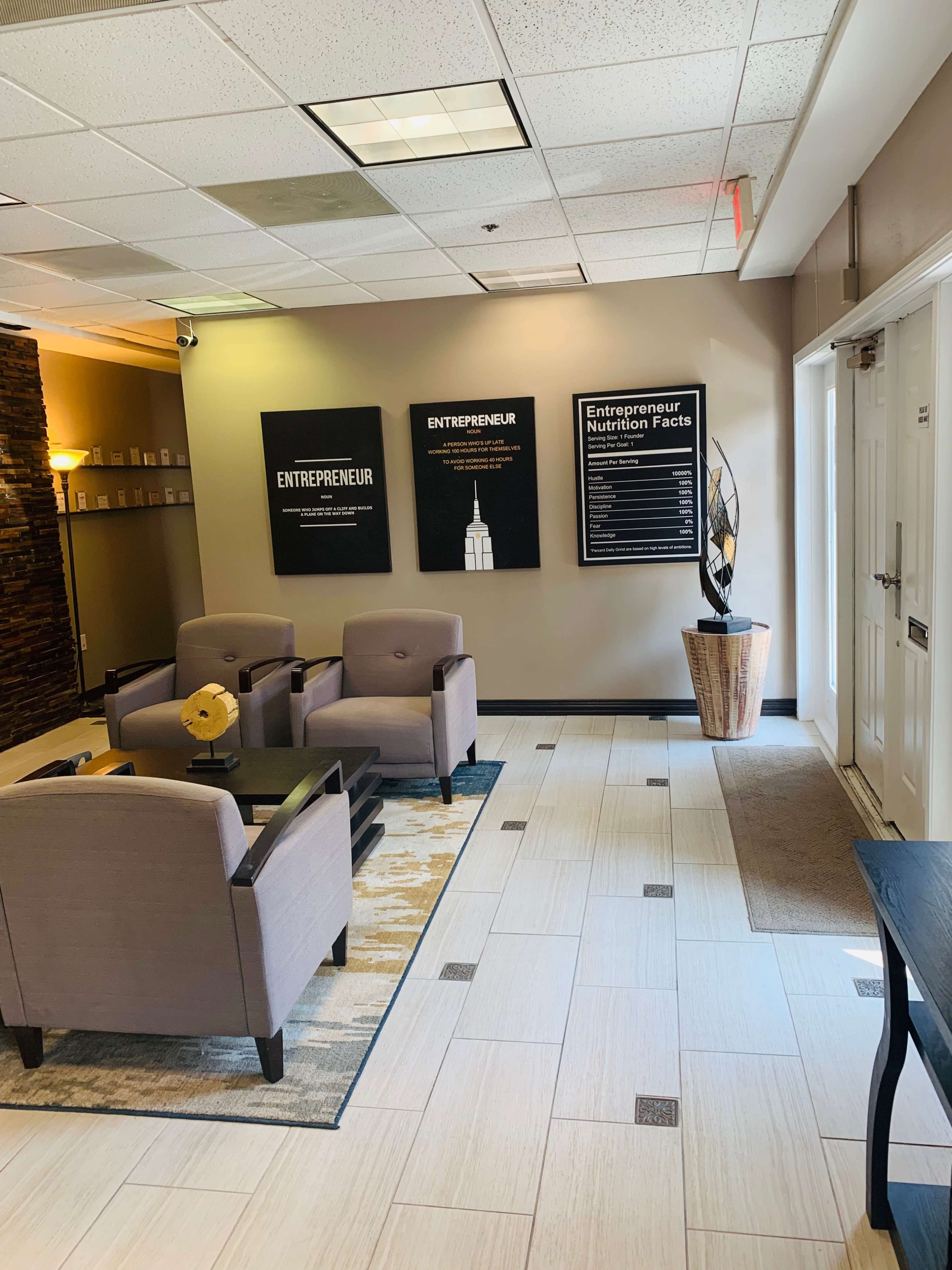 The image shows a modern waiting area with gray upholstered chairs, a small table, and wall art featuring the word "ENTREPRENEUR" alongside informational displays.