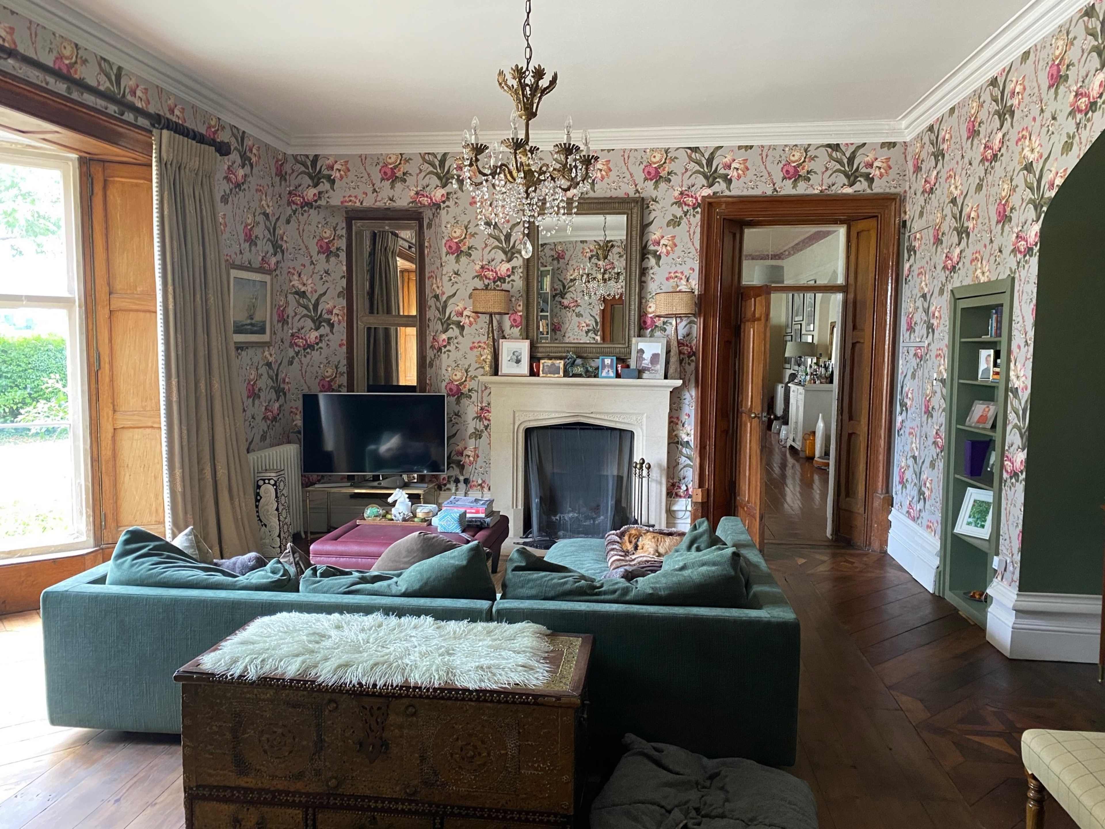 The living room features a floral wallpaper, a chandelier, a central fireplace, and a sectional sofa arranged around a coffee table.