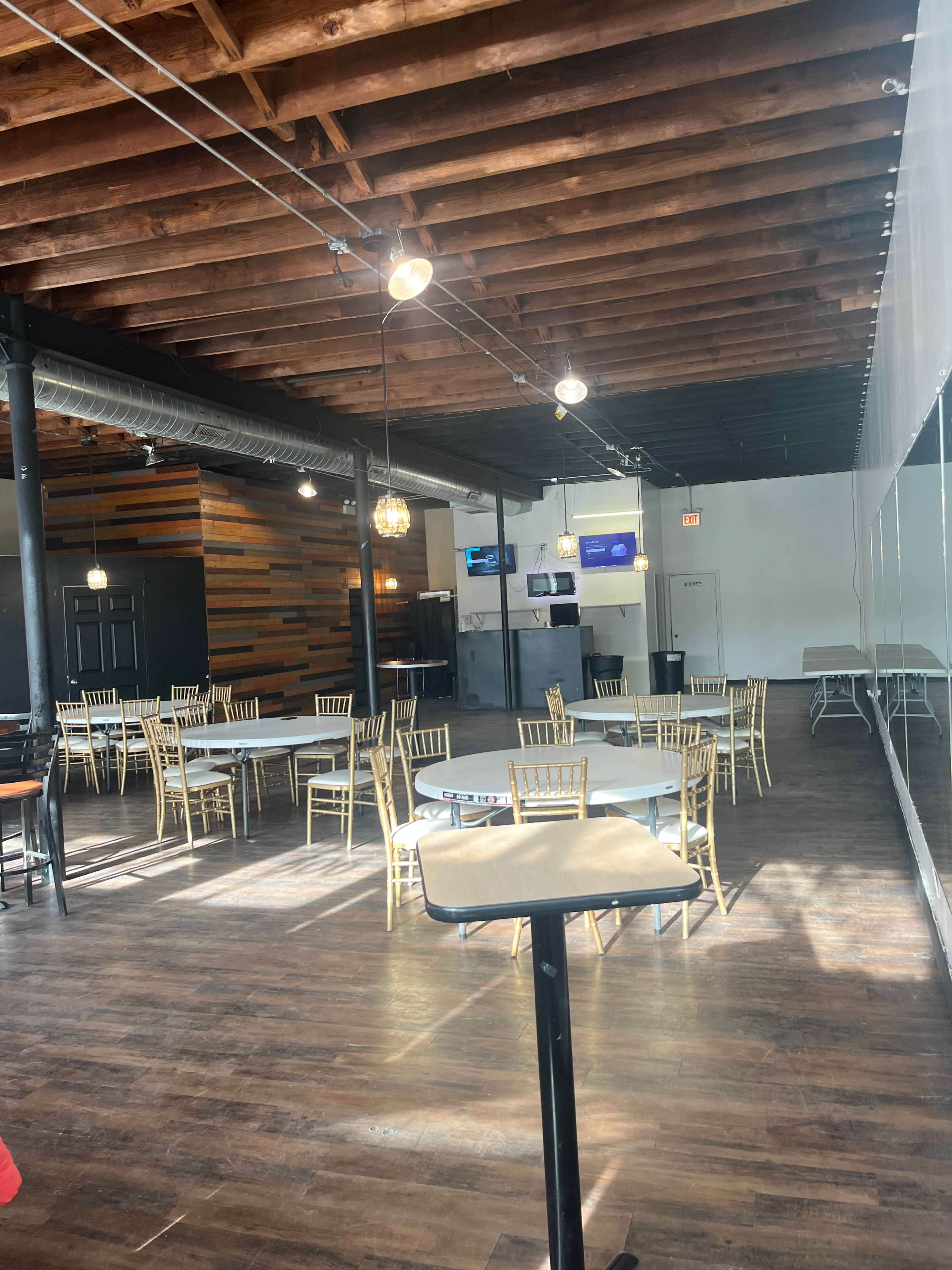 The image shows a spacious, well-lit restaurant interior with wooden beams overhead, several round tables surrounded by gold chairs, and a bar counter in the foreground.