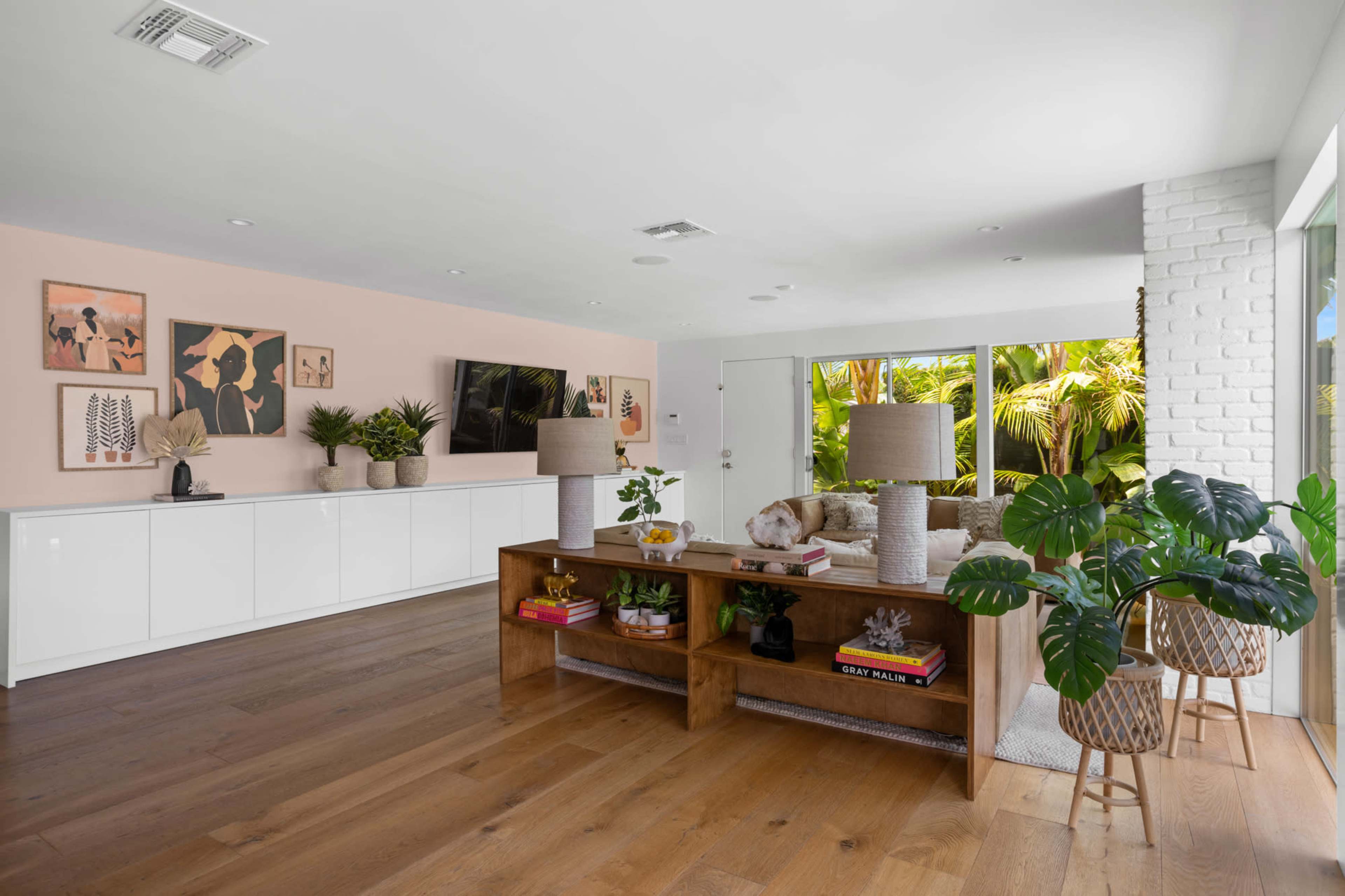 A modern living room features a wooden TV console with lamps, plants, and decor against a pale pink wall, with a view of greenery through large windows.
