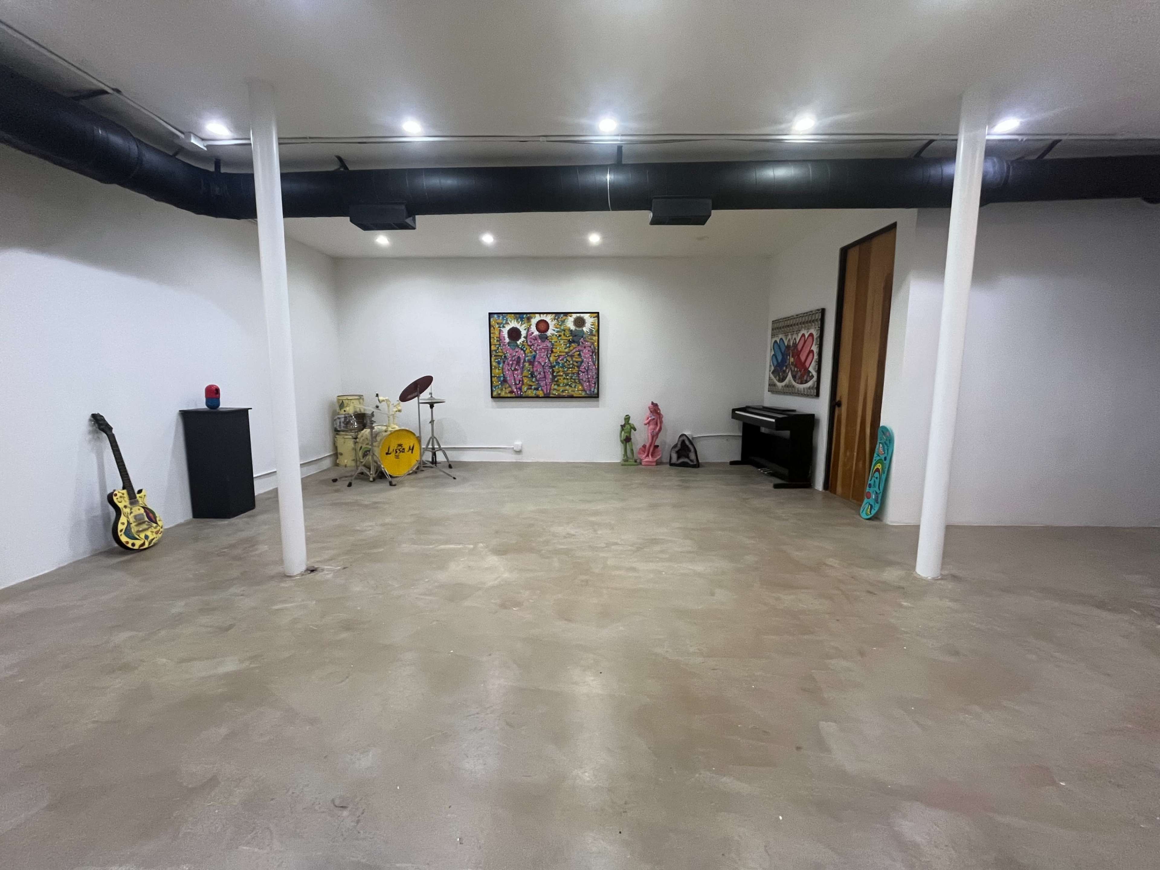 The image shows a spacious, well-lit basement featuring a drum set, a piano, and various art pieces along with musical instruments and decorative objects.