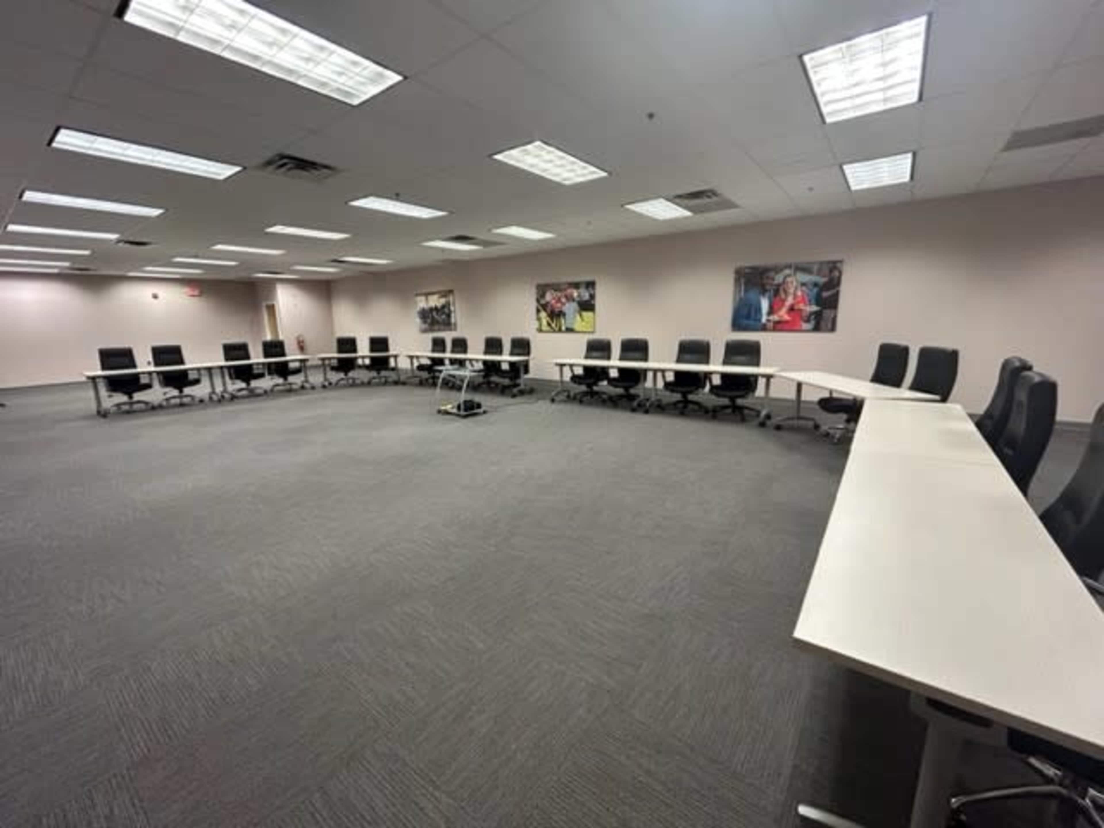 The image shows a spacious conference room arranged with a large oval table and rows of black chairs around it, illuminated by ceiling lights.