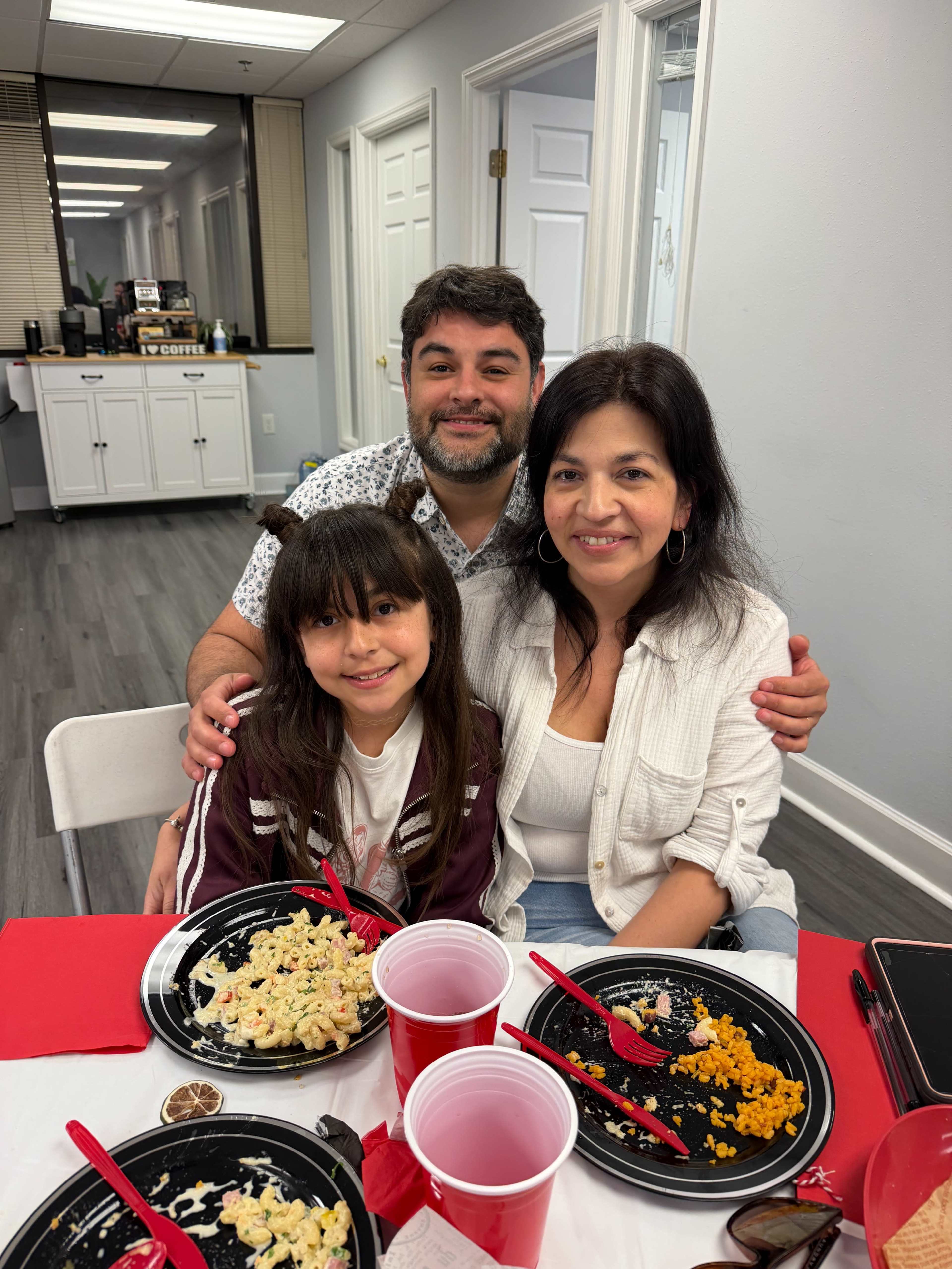 A family of three sits together at a table with half-eaten plates of food and red cups, smiling at the camera.