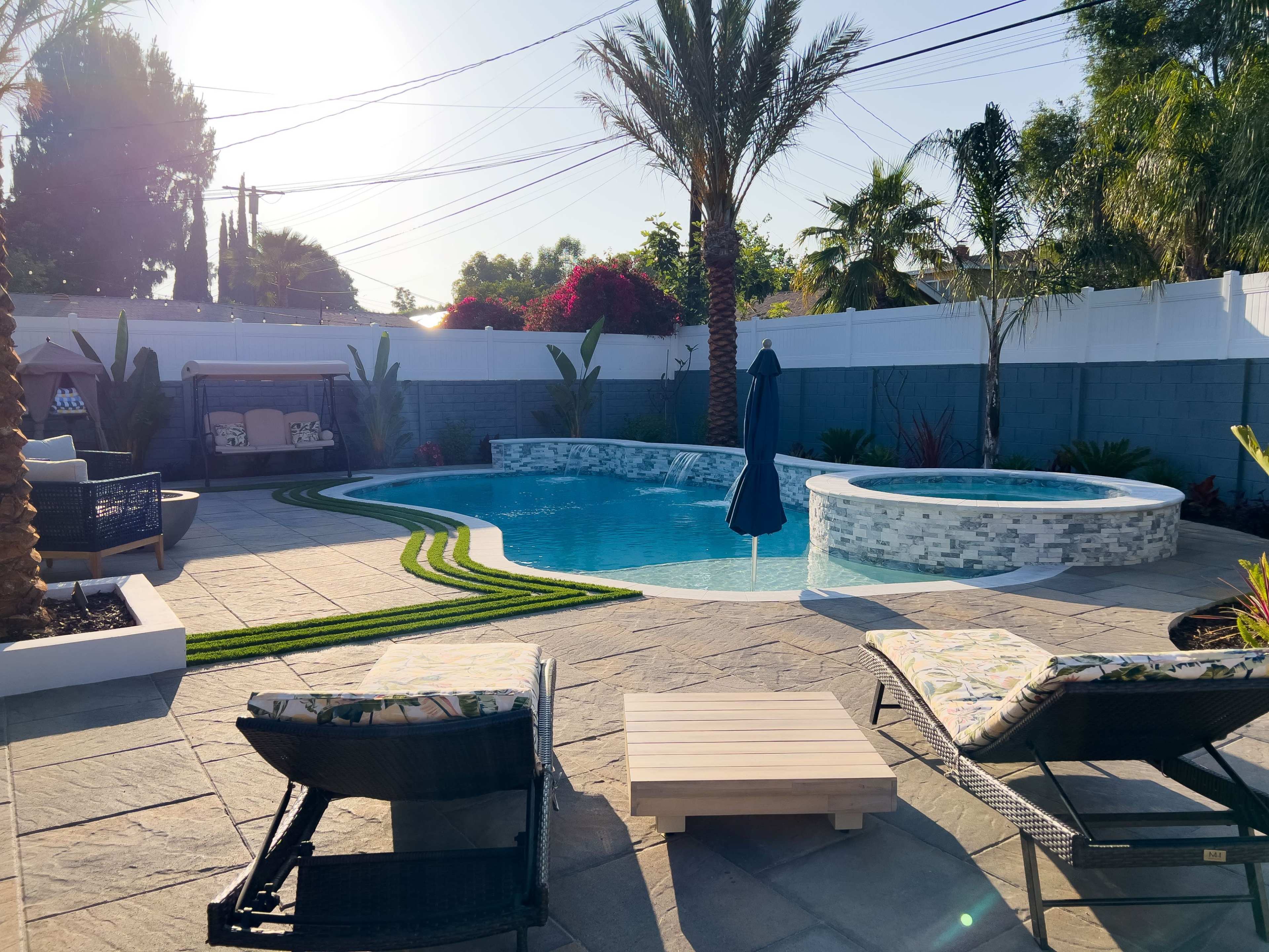 A swimming pool with a spa surrounded by landscaped palms and stone flooring, along with lounge chairs and a pergola in a residential backyard.