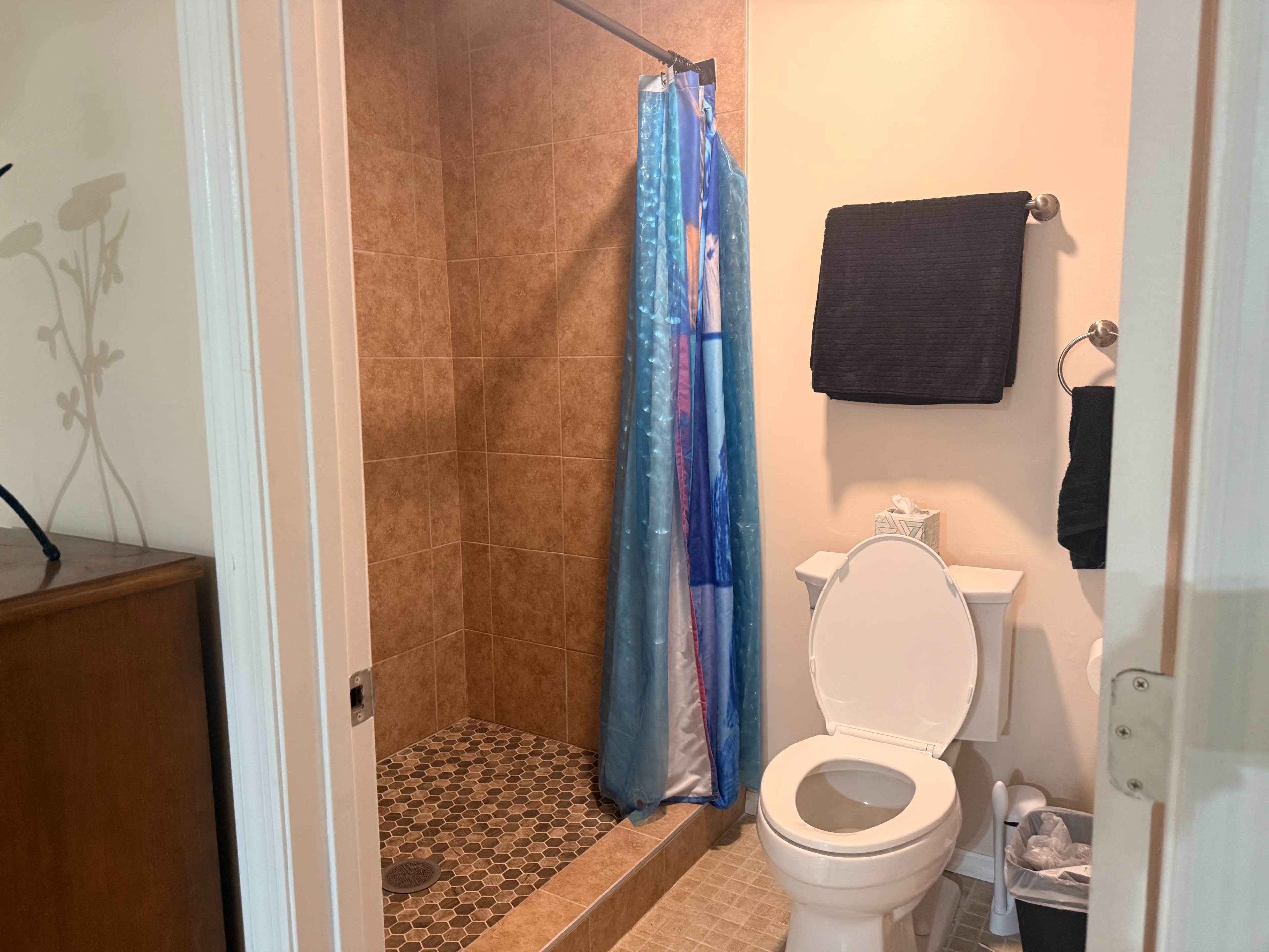 The image shows a small bathroom featuring a shower with a blue curtain, a toilet, and a tiled floor.