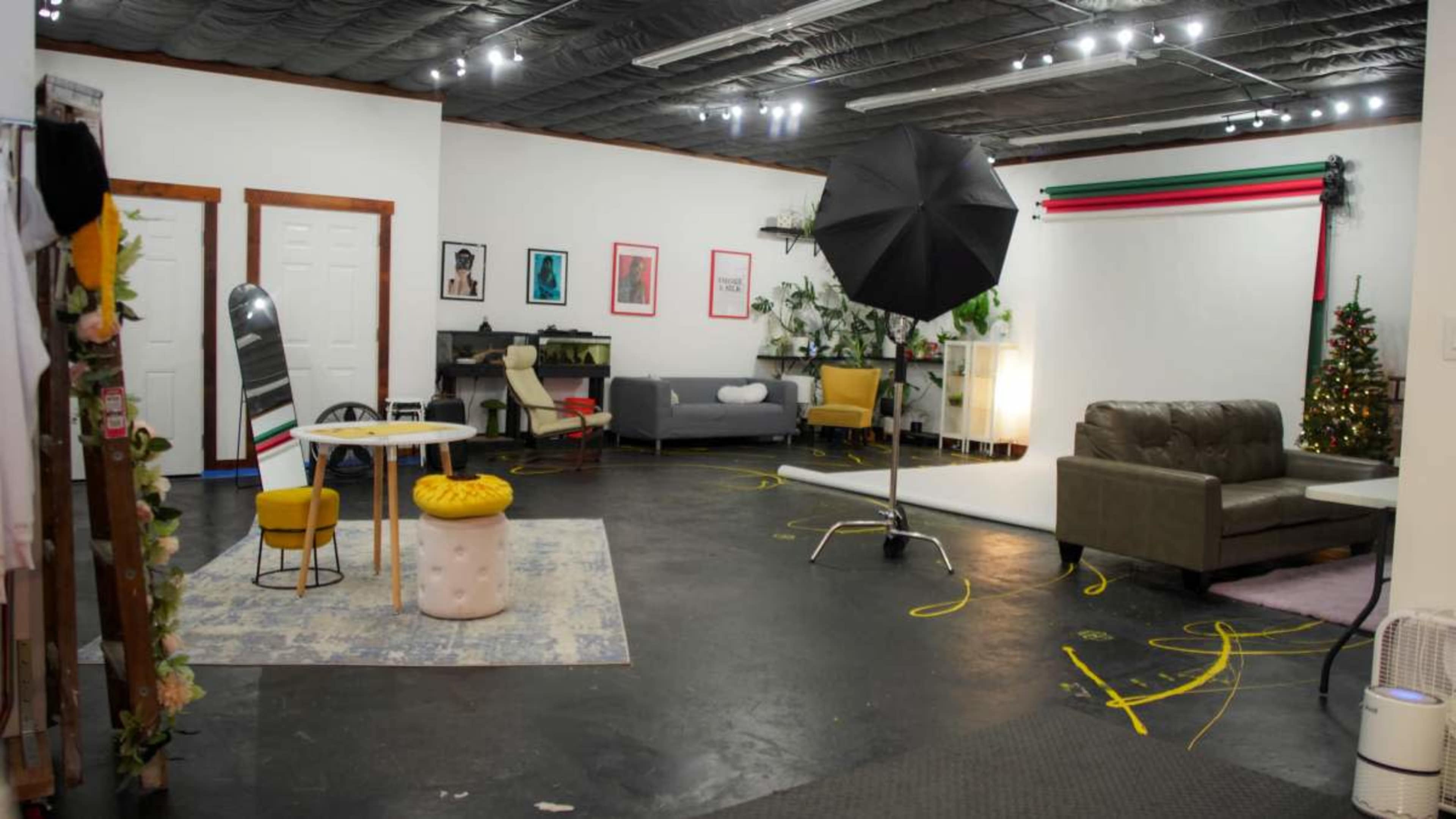 The image shows a well-lit studio space with various styled areas, including a gray sofa, a yellow chair, a circular table, and photography equipment set up for a shoot.