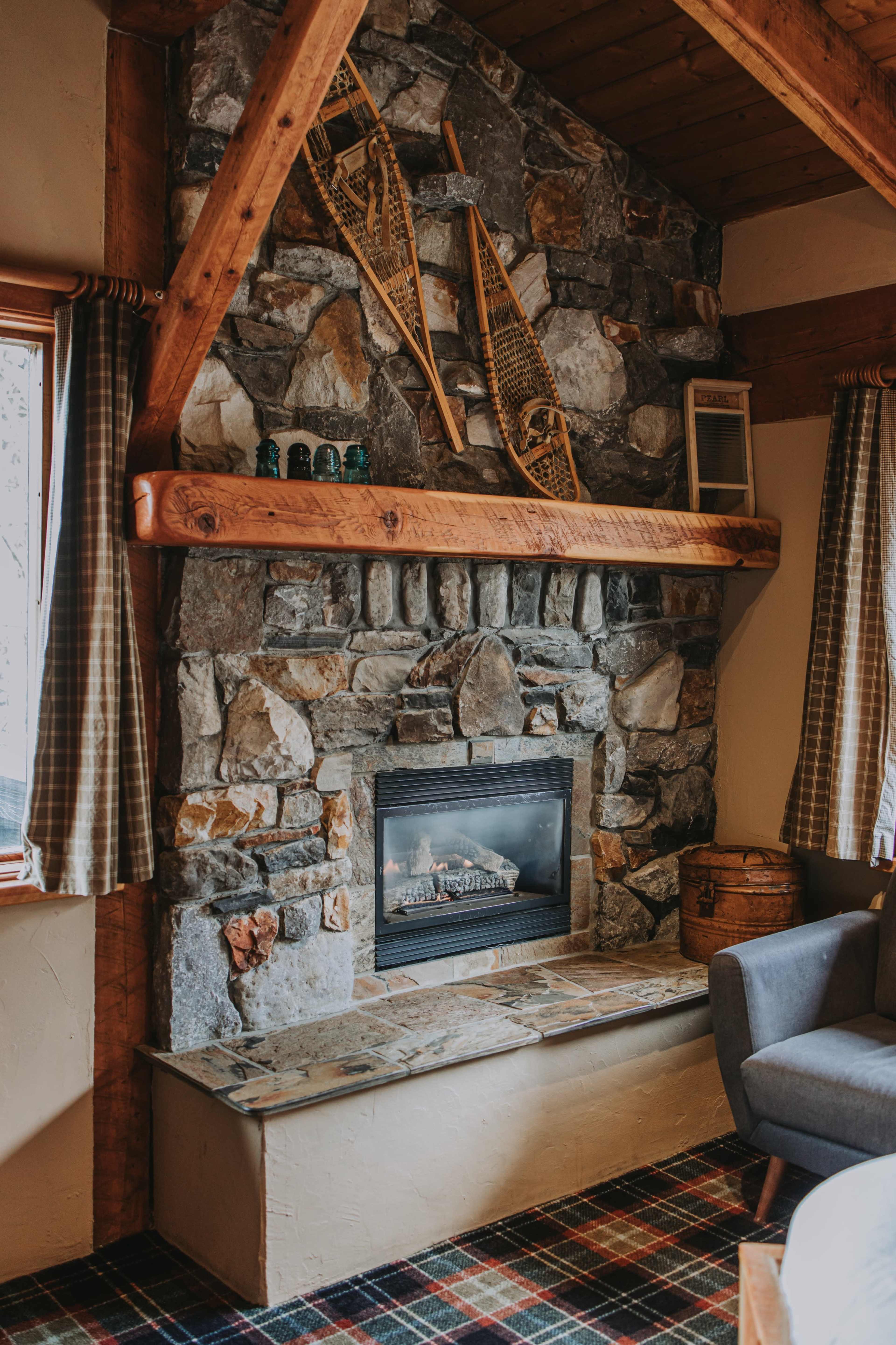 The image shows a cozy stone fireplace with wooden beams above and decoratively arranged snowshoes on the wall, surrounded by rustic furnishings and patterned curtains.