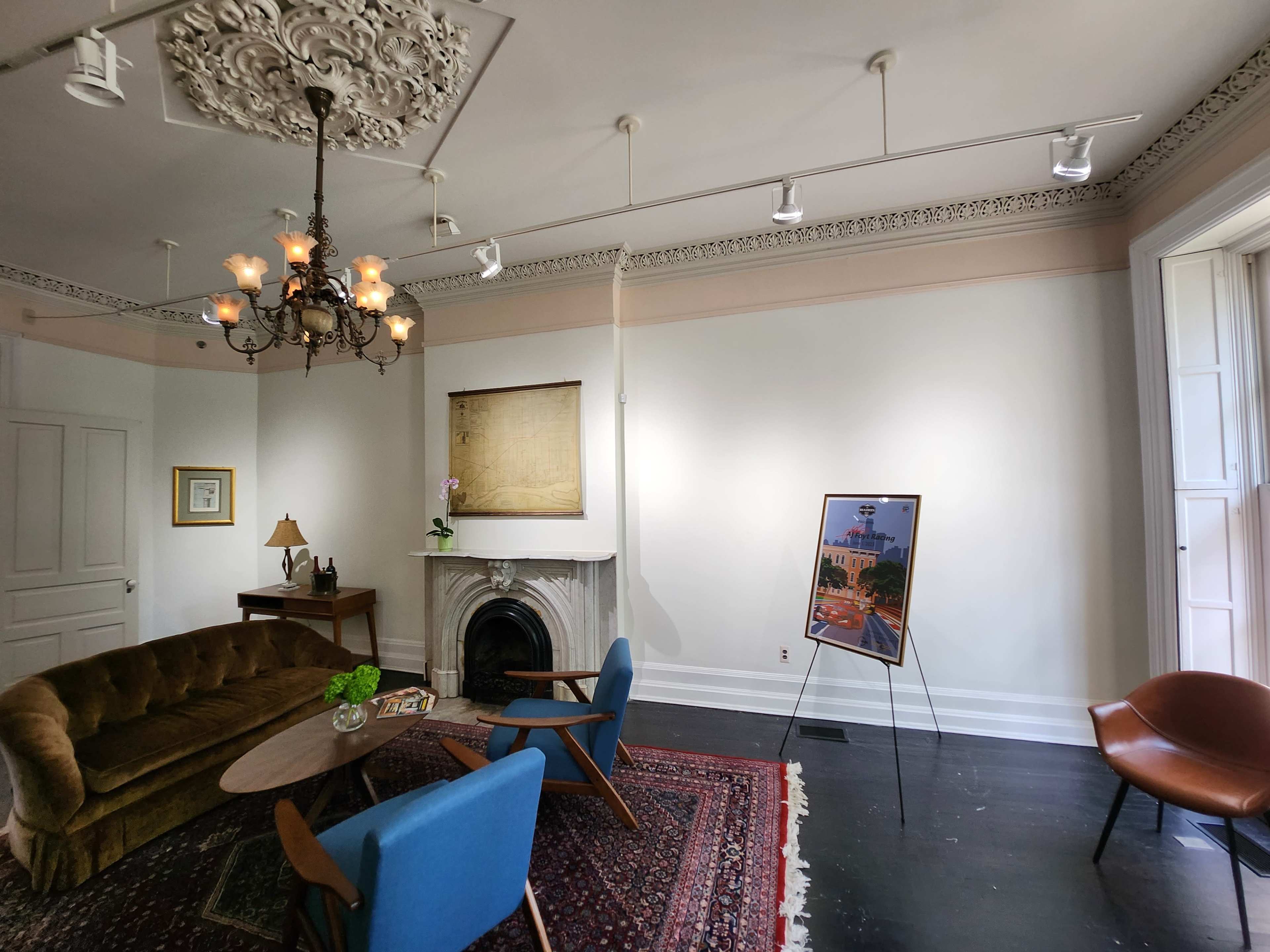 A well-lit, elegantly furnished room featuring a vintage brown sofa, blue chairs, a round table, and a fireplace, with artwork displayed on the walls.