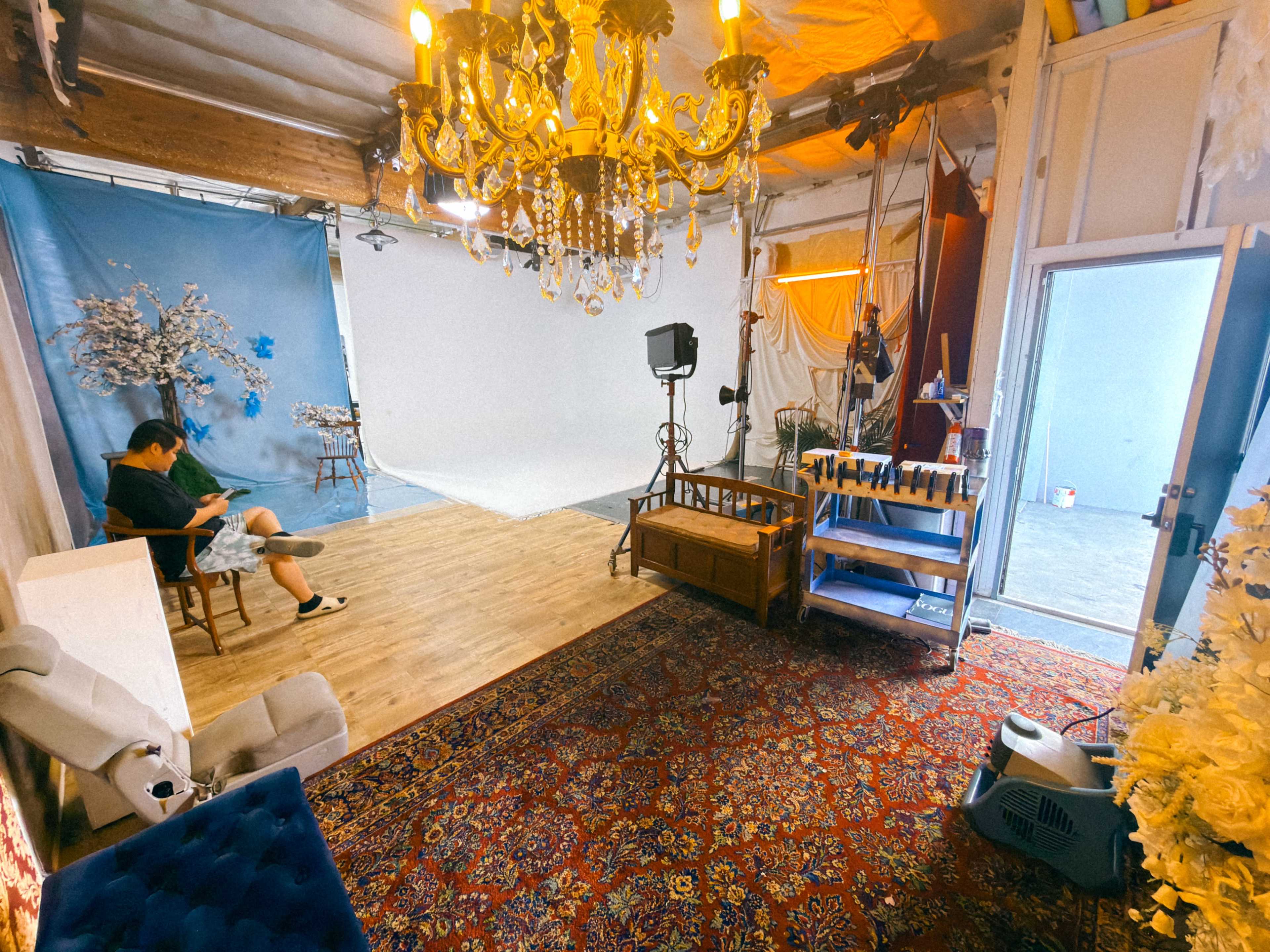 The image shows a photography studio with a wooden floor, decorative elements including a chandelier and a rug, and a person sitting while using a phone.