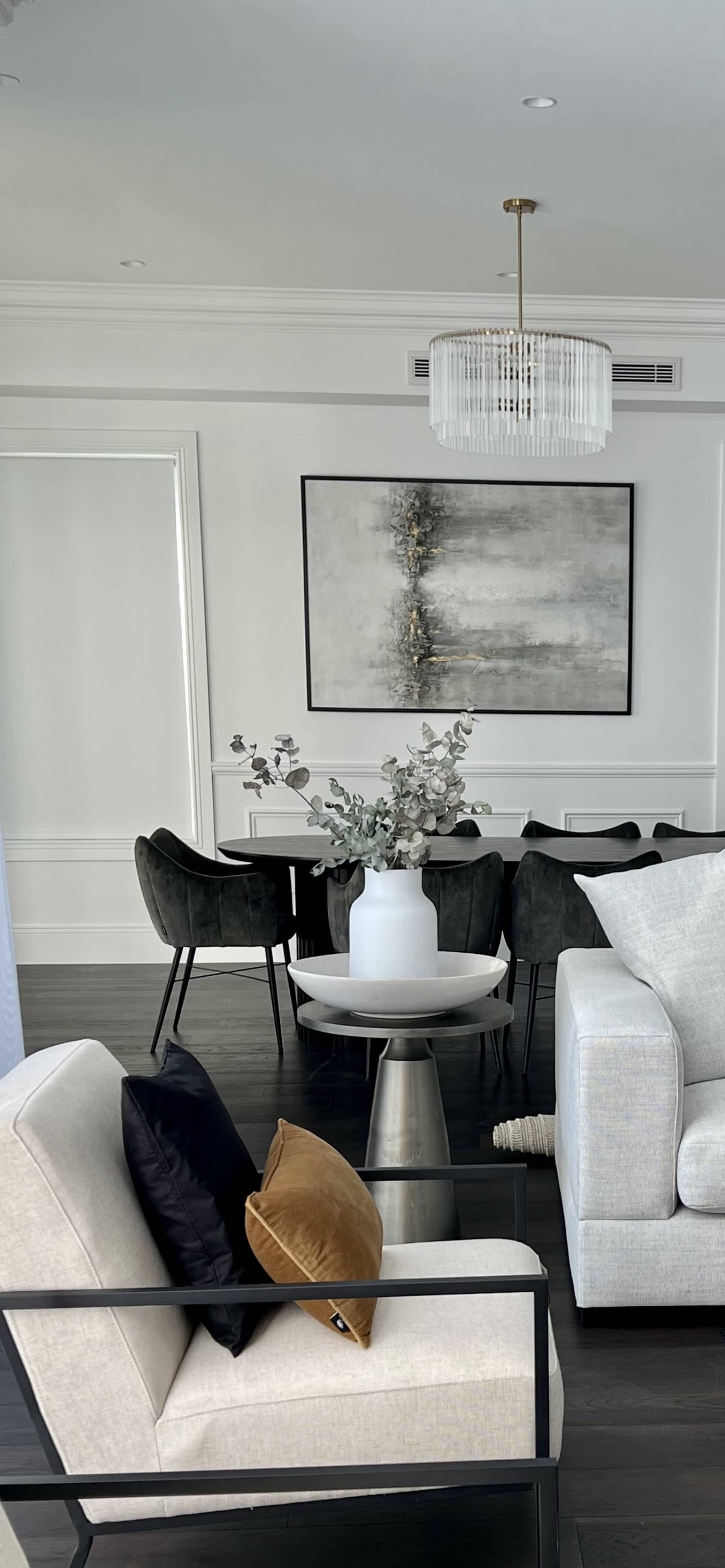 The image shows a modern, well-lit living room and dining area featuring a light-colored sofa, a round coffee table with a vase of flowers, and a dining table in the background surrounded by dark chairs.