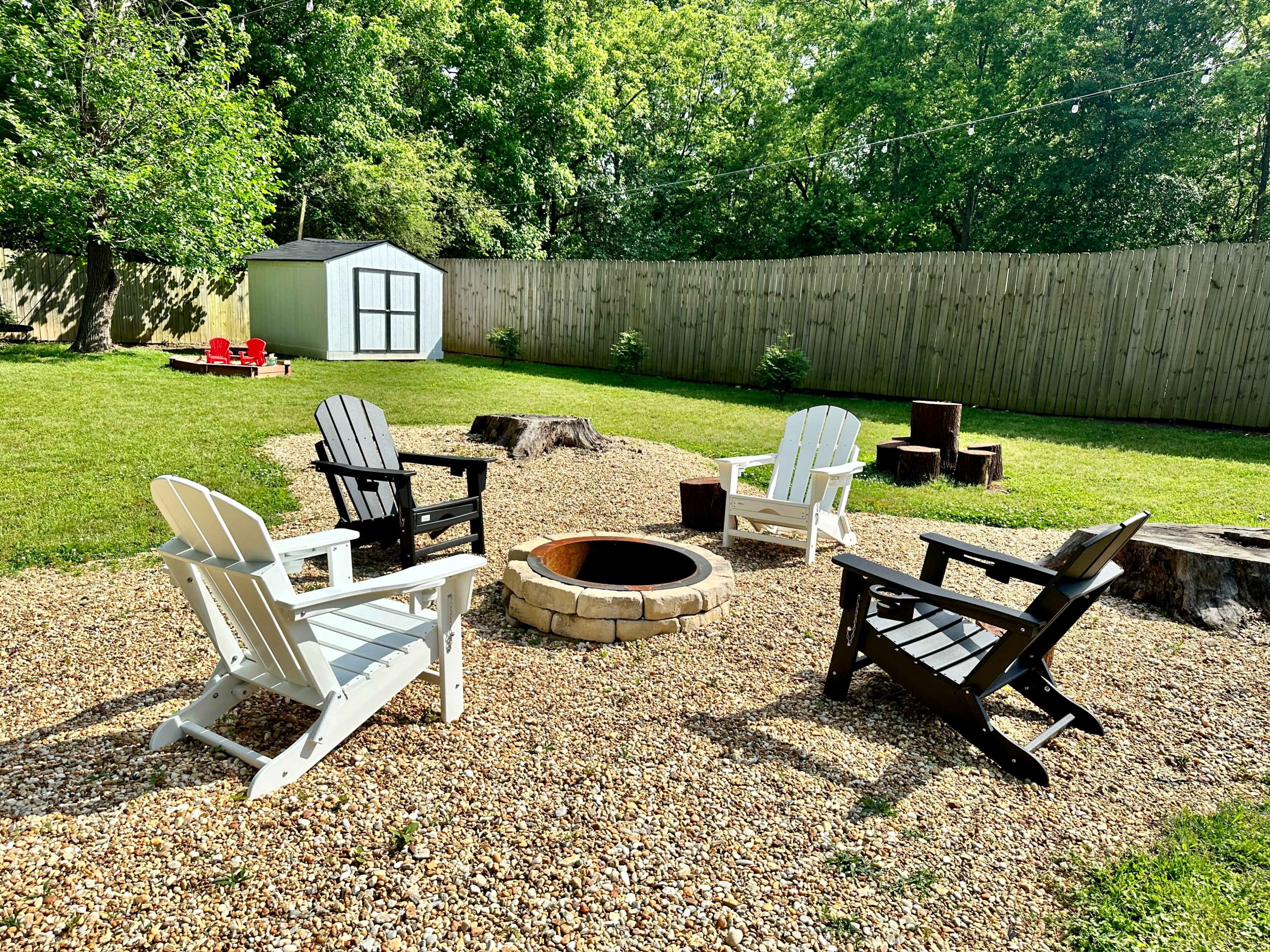 The image shows a fire pit surrounded by four outdoor chairs on a gravel area in a grassy backyard, with a wooden fence and a storage shed in the background.