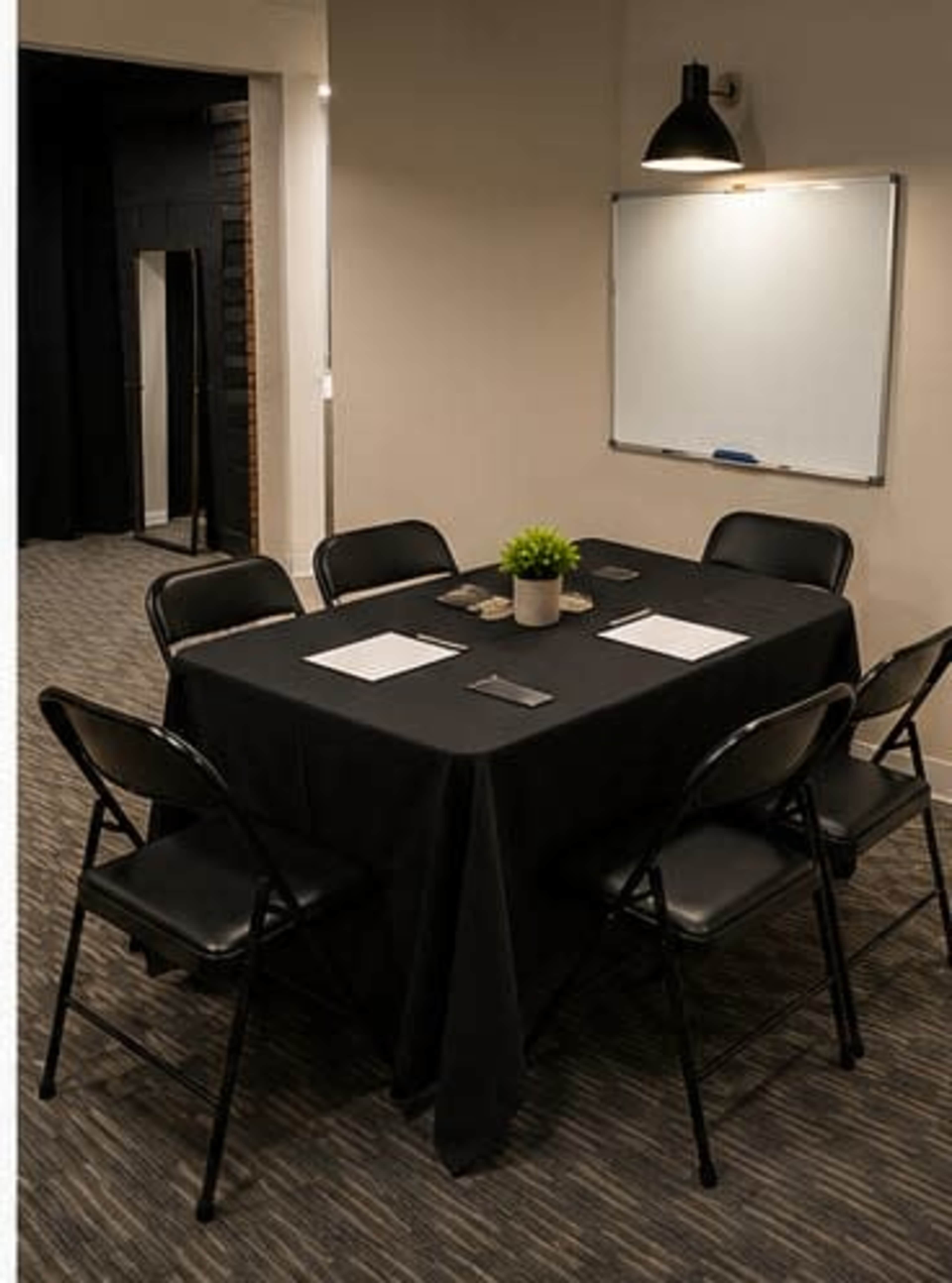 A conference room features a black table surrounded by six black folding chairs, with a potted plant in the center and a whiteboard on the wall.
