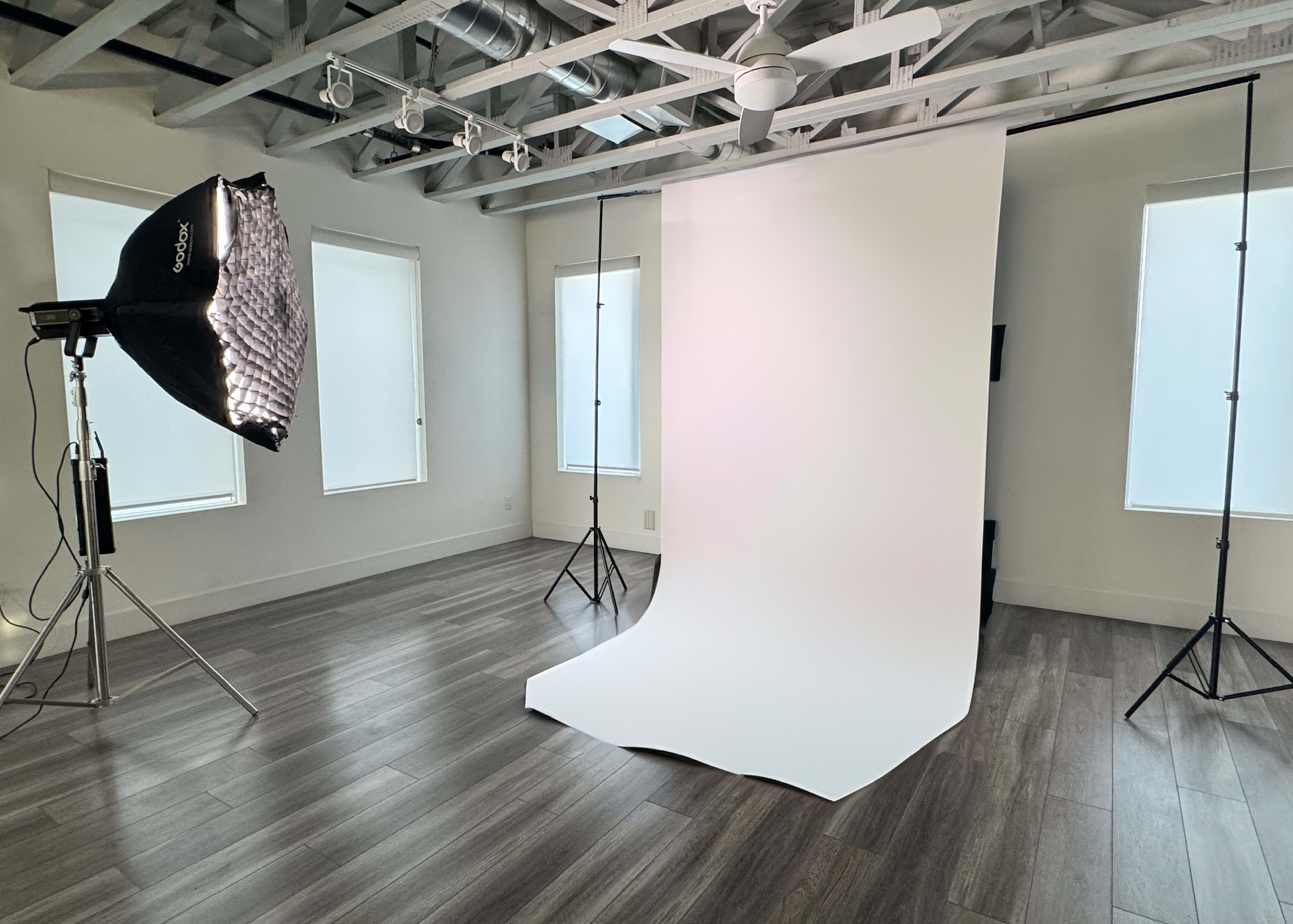 The image shows a photography studio with a large softbox light and a seamless white backdrop, set against a wooden floor and bright windows.