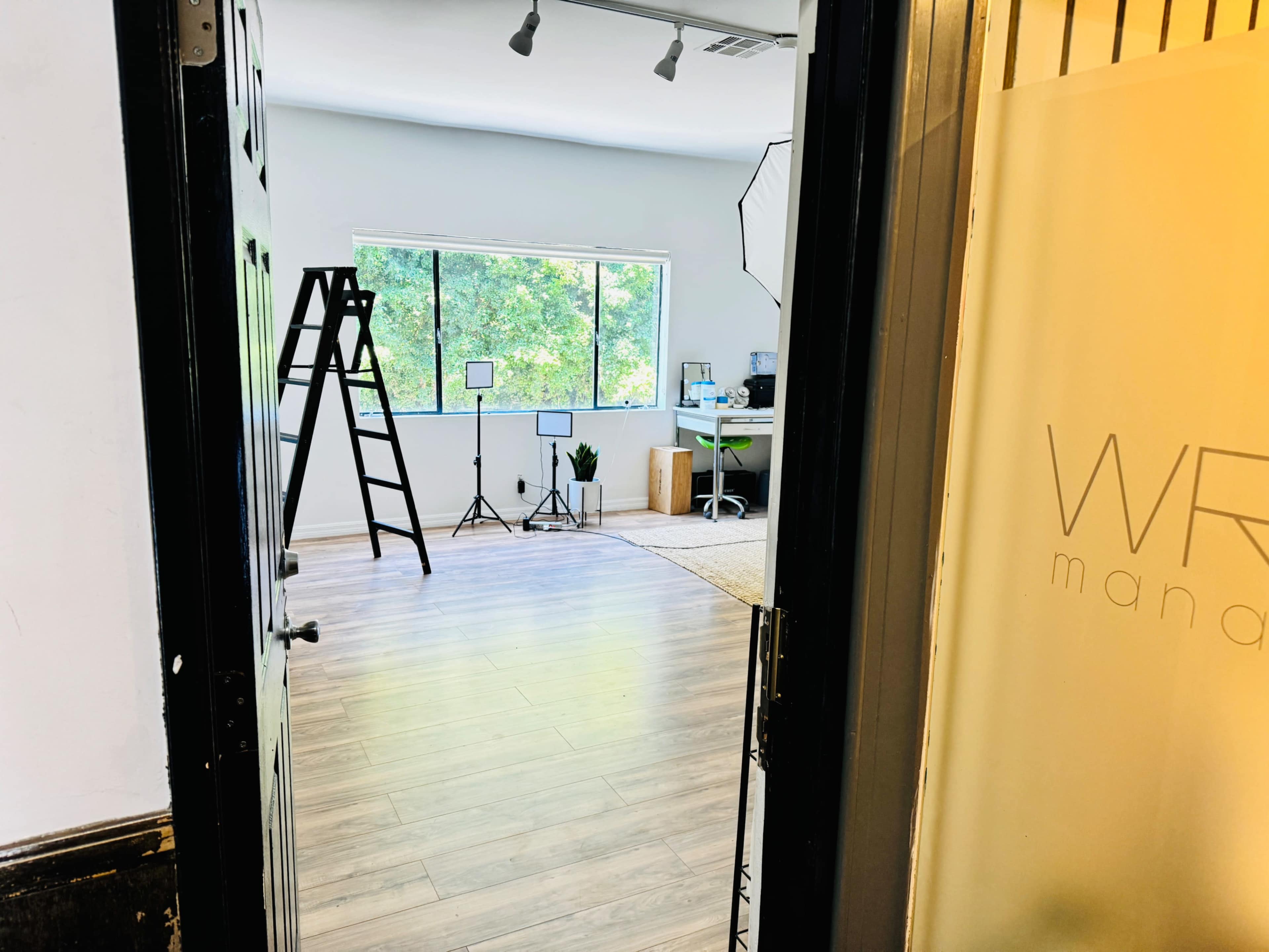 An empty room with large windows, a ladder, a plant, and a desk setup in the corner.