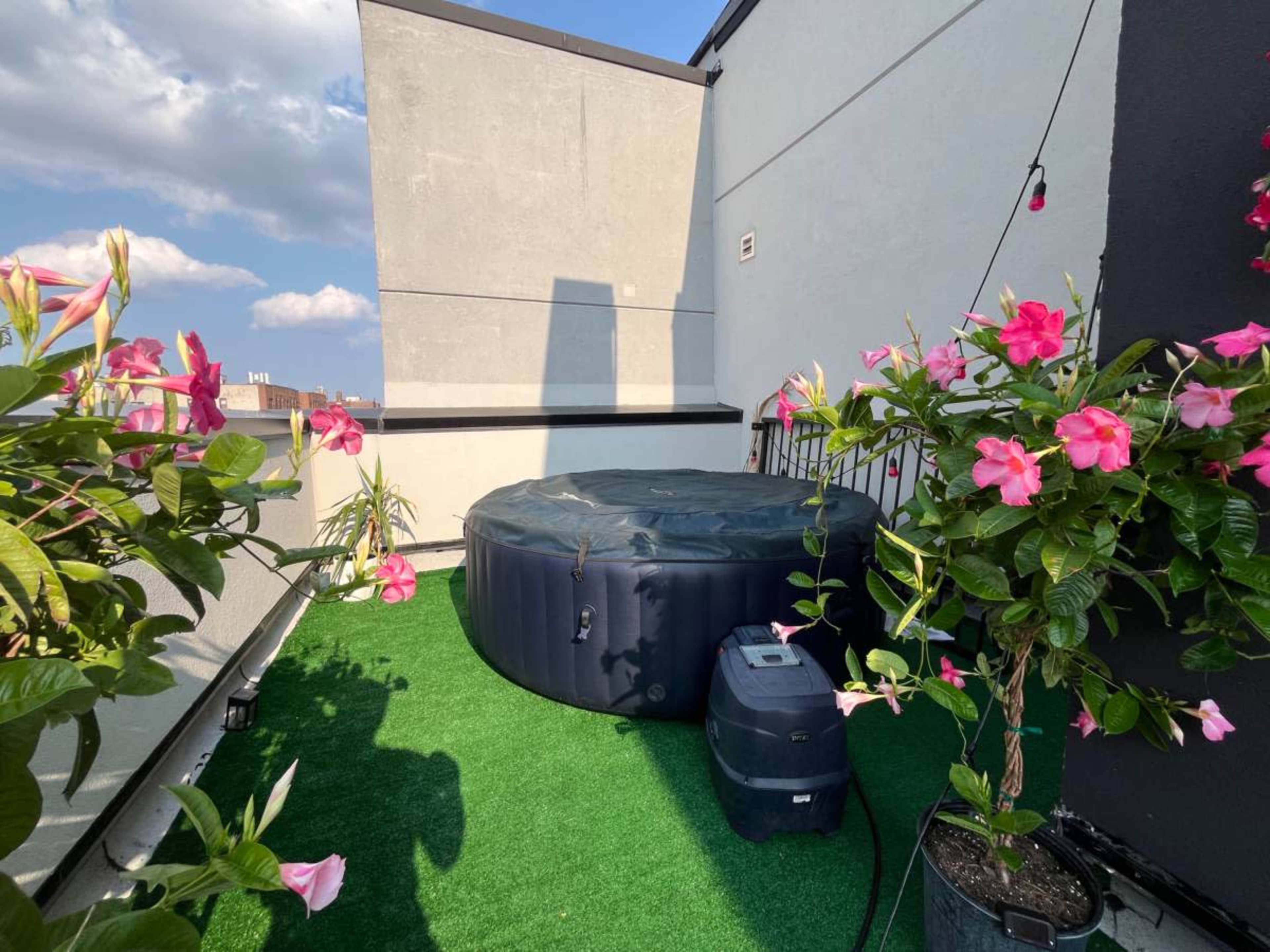 A hot tub covered with a black tarp is placed on a green synthetic lawn surrounded by flowering plants on a rooftop terrace.
