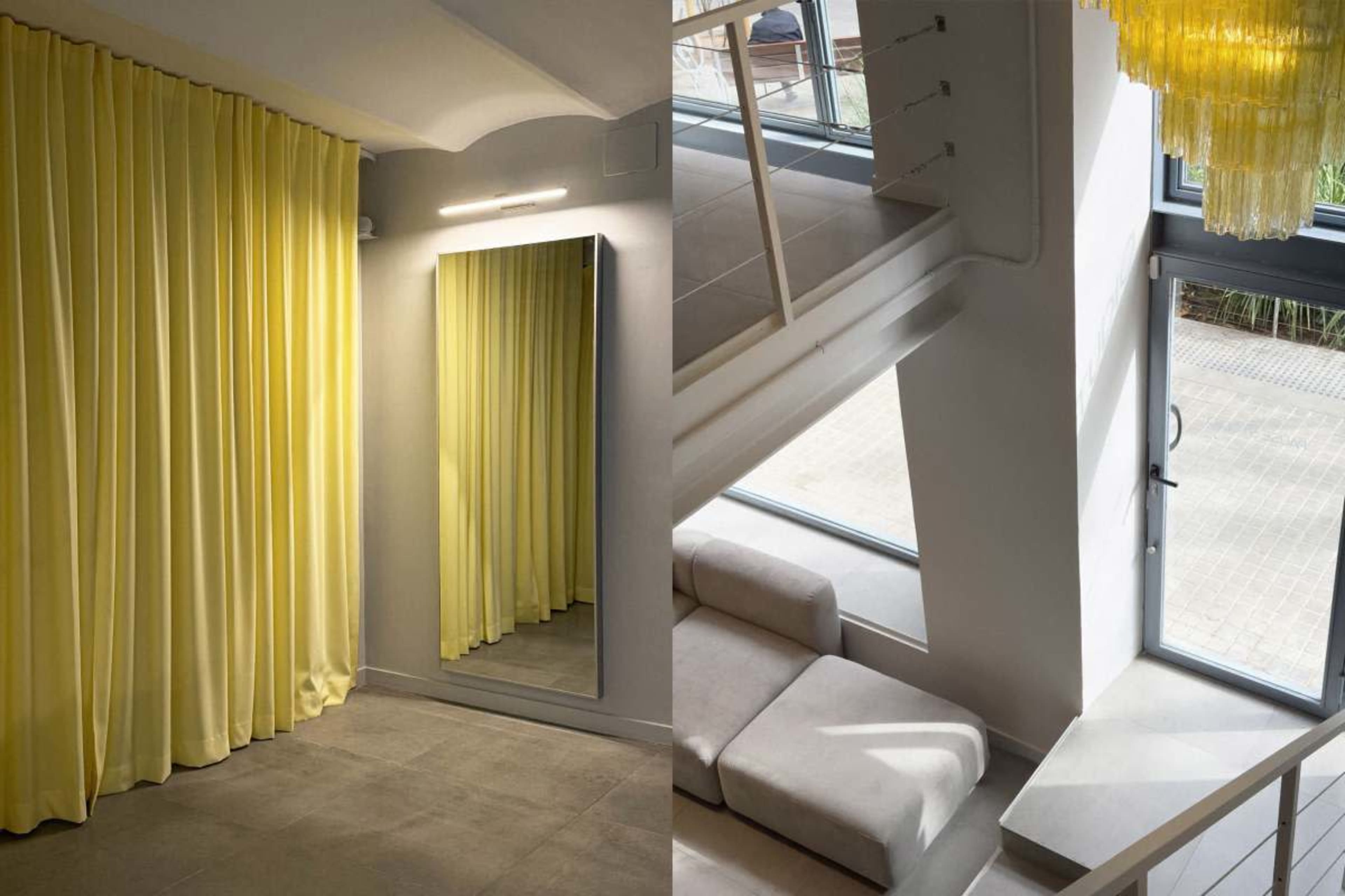 The image shows a modern interior space featuring yellow curtains and a mirror on one side, and a light-filled area with a staircase and large window on the other.