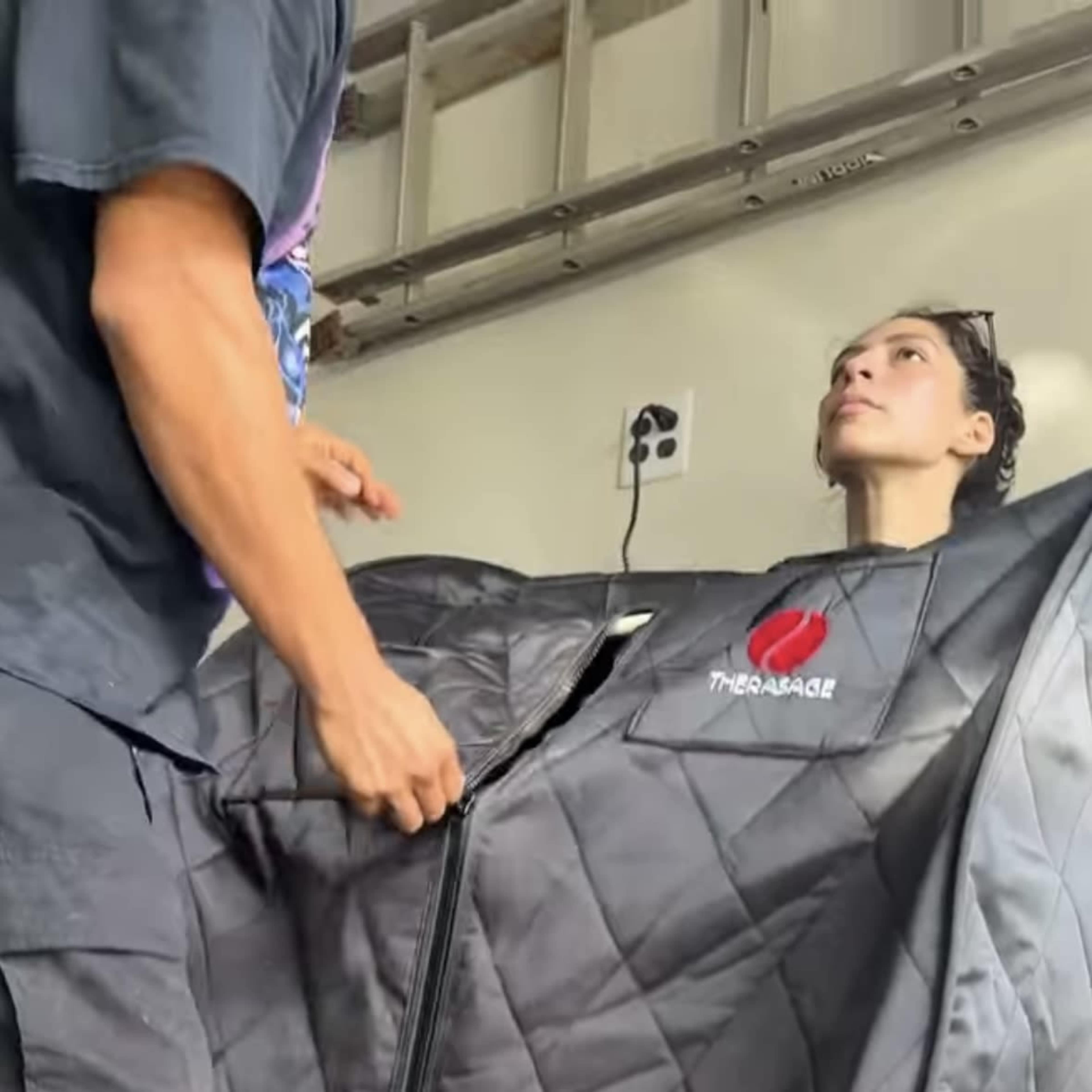 A person stands facing a seated individual who is inside a zippered thermal suit.