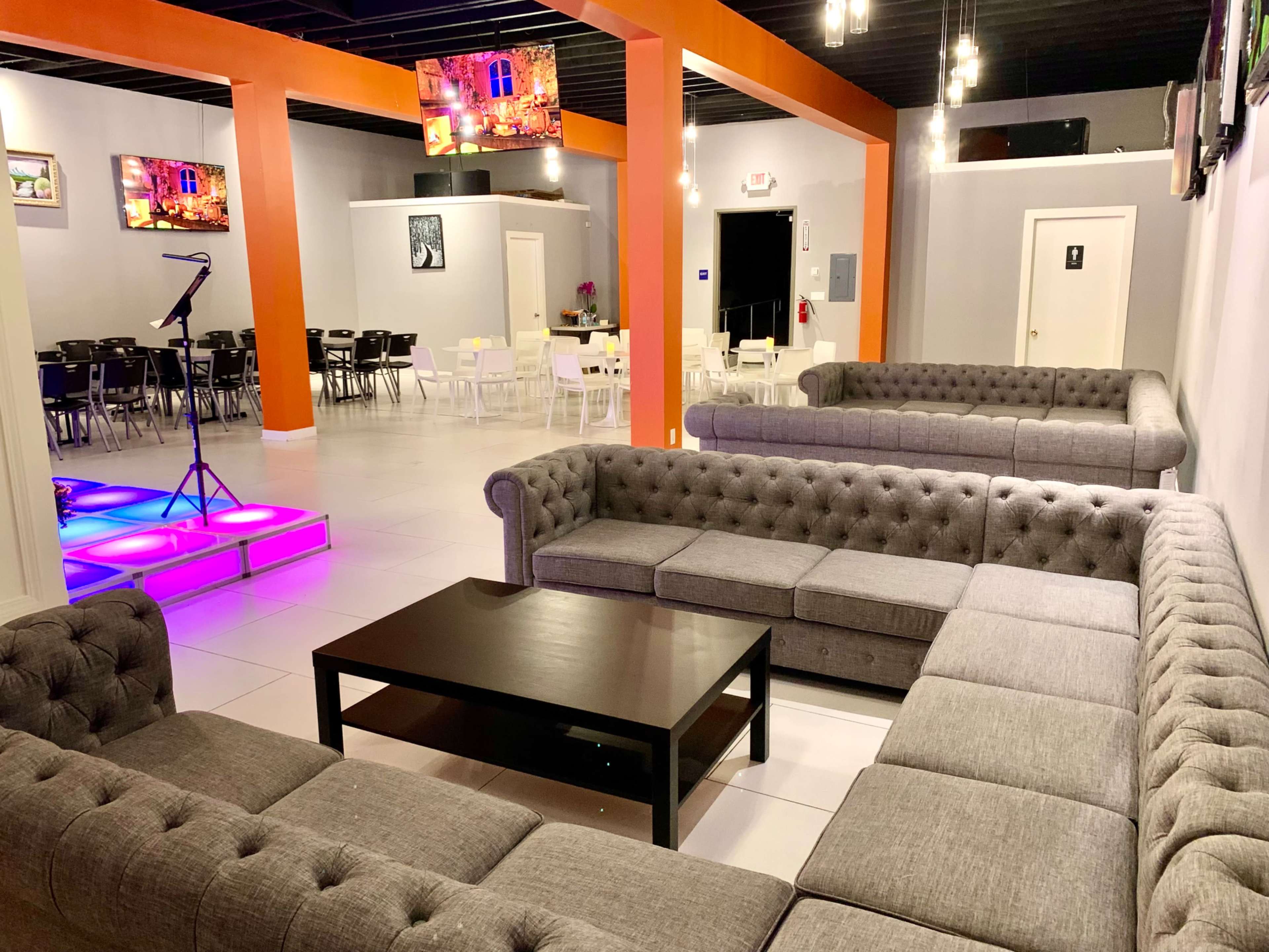 The image shows a modern interior featuring gray sectional sofas arranged around a black coffee table, with colorful lighting and multiple televisions on the walls.