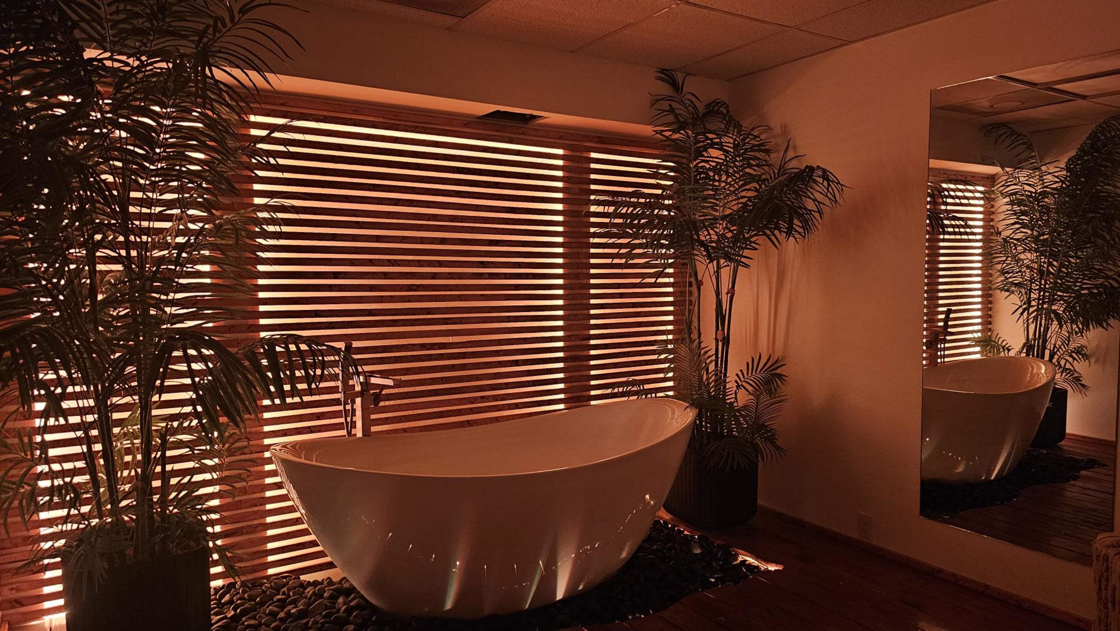 A freestanding bathtub is positioned in a room with wooden slatted blinds and potted plants, creating a serene ambiance.