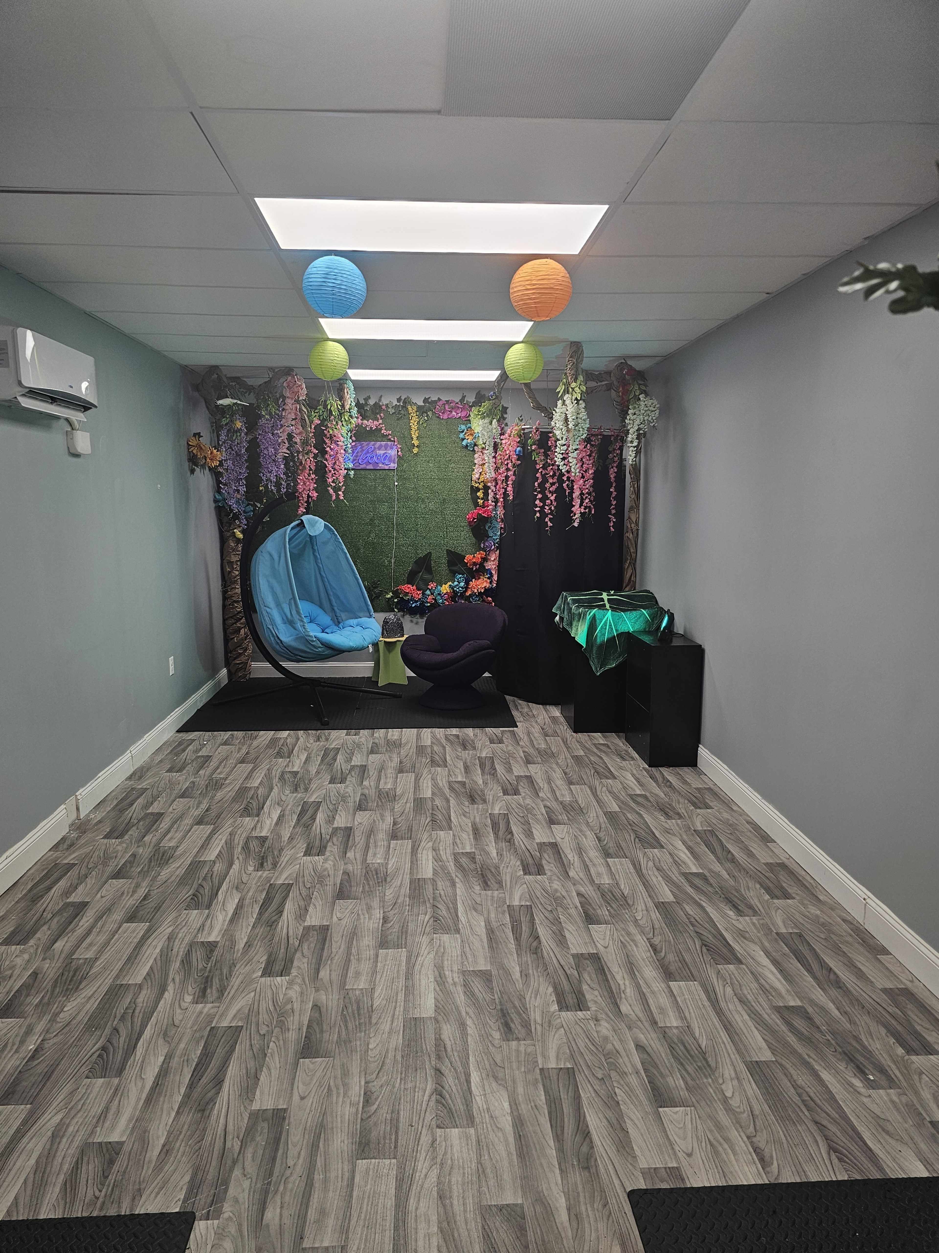 A spacious room features a blue hammock chair, decorative plants, and colorful lanterns hanging from the ceiling, with a green backdrop and wooden flooring.