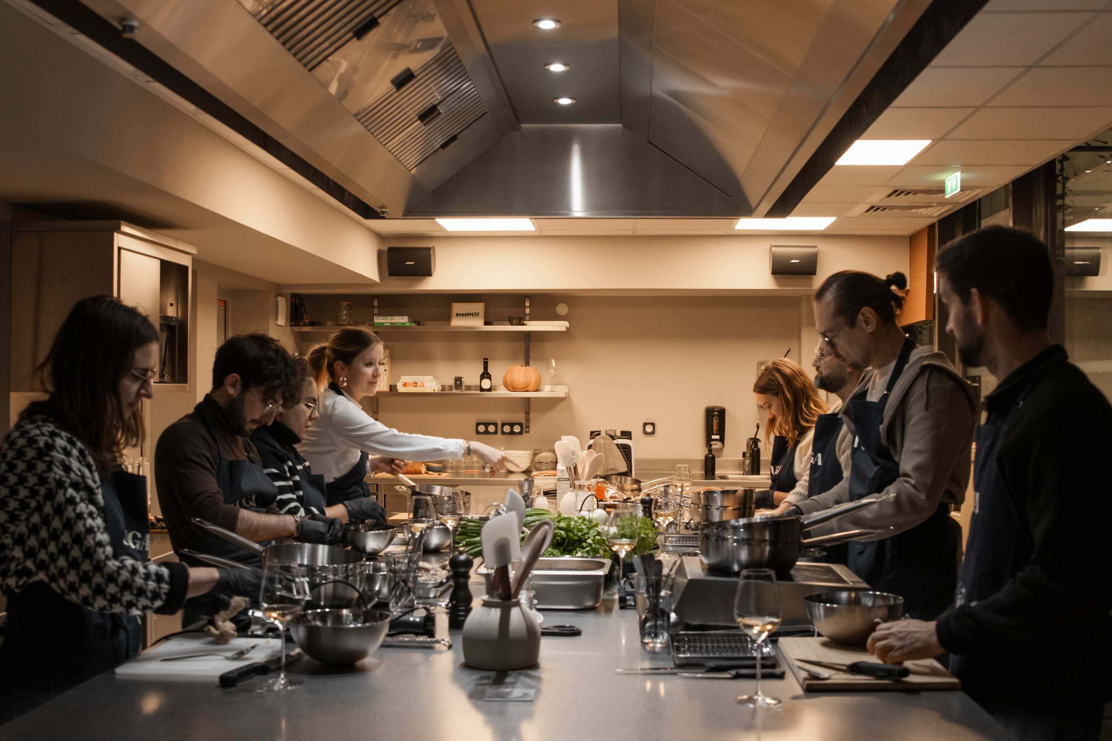 A group of people in aprons collaborate in a well-equipped kitchen, preparing various dishes at cooking stations.