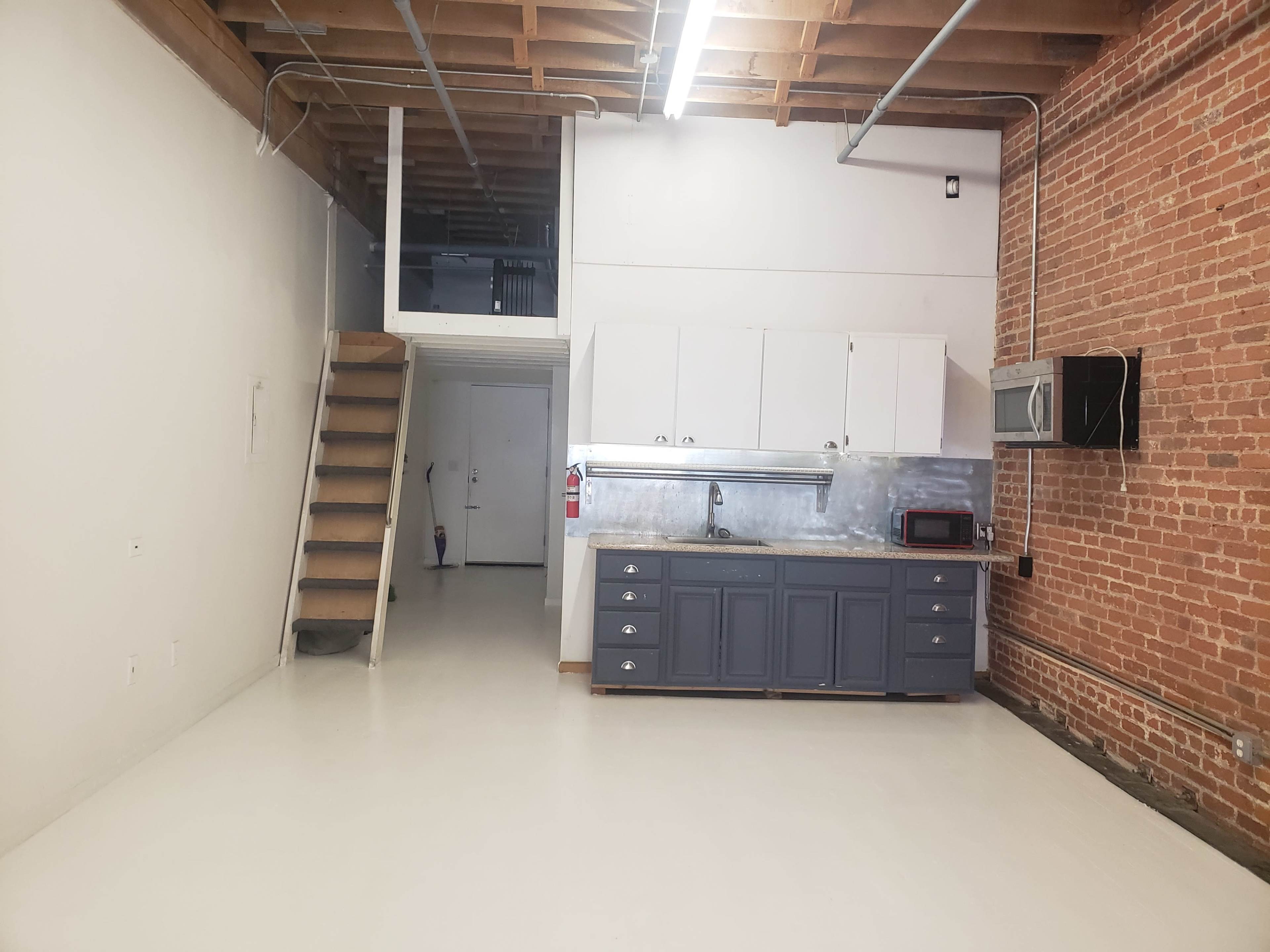 The image shows an open-concept space featuring a kitchen area with gray cabinetry, a staircase leading to a loft, and exposed brick walls.