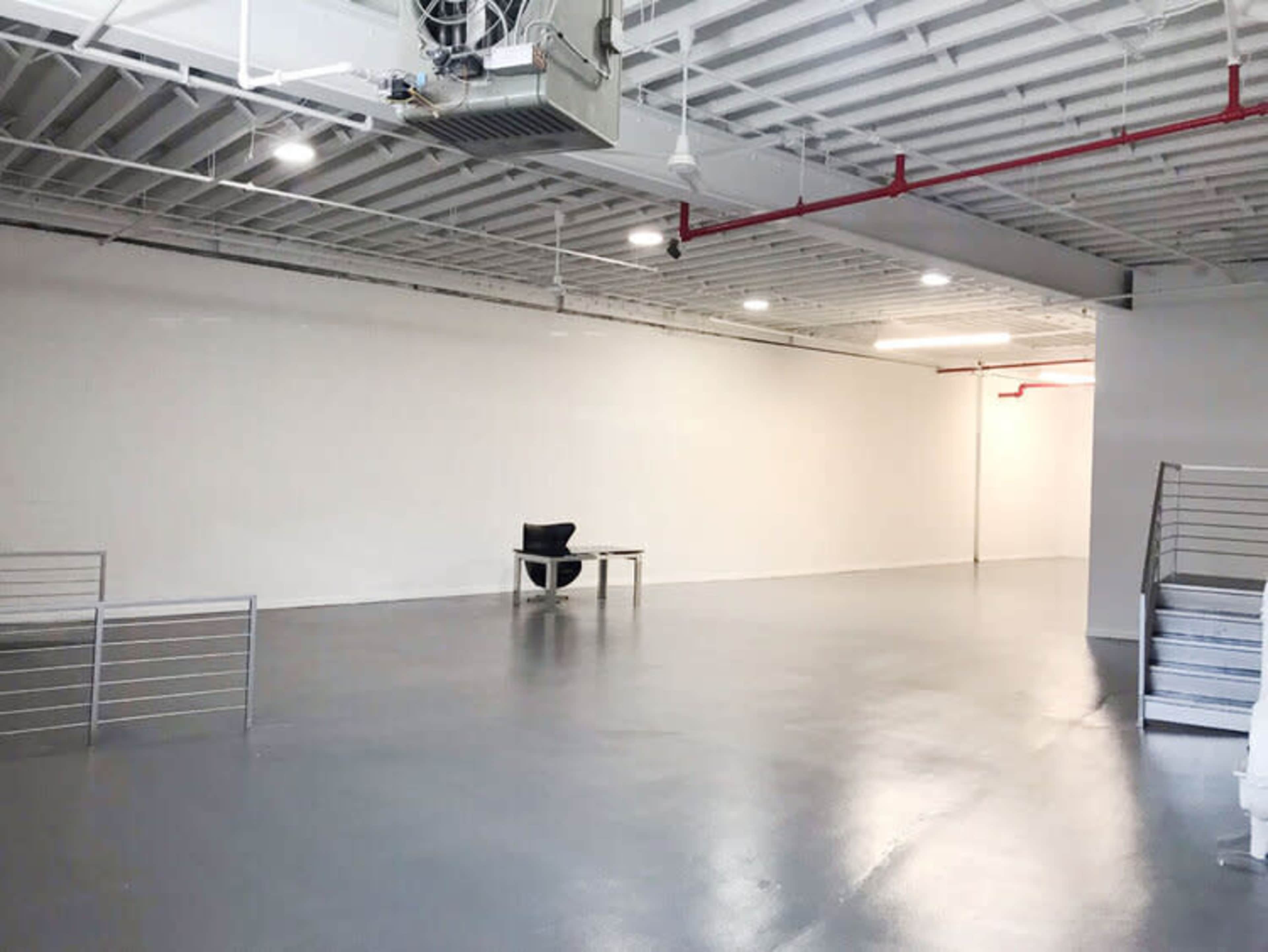 A large, empty room with a polished floor, featuring a single black chair and a table against a plain white wall.