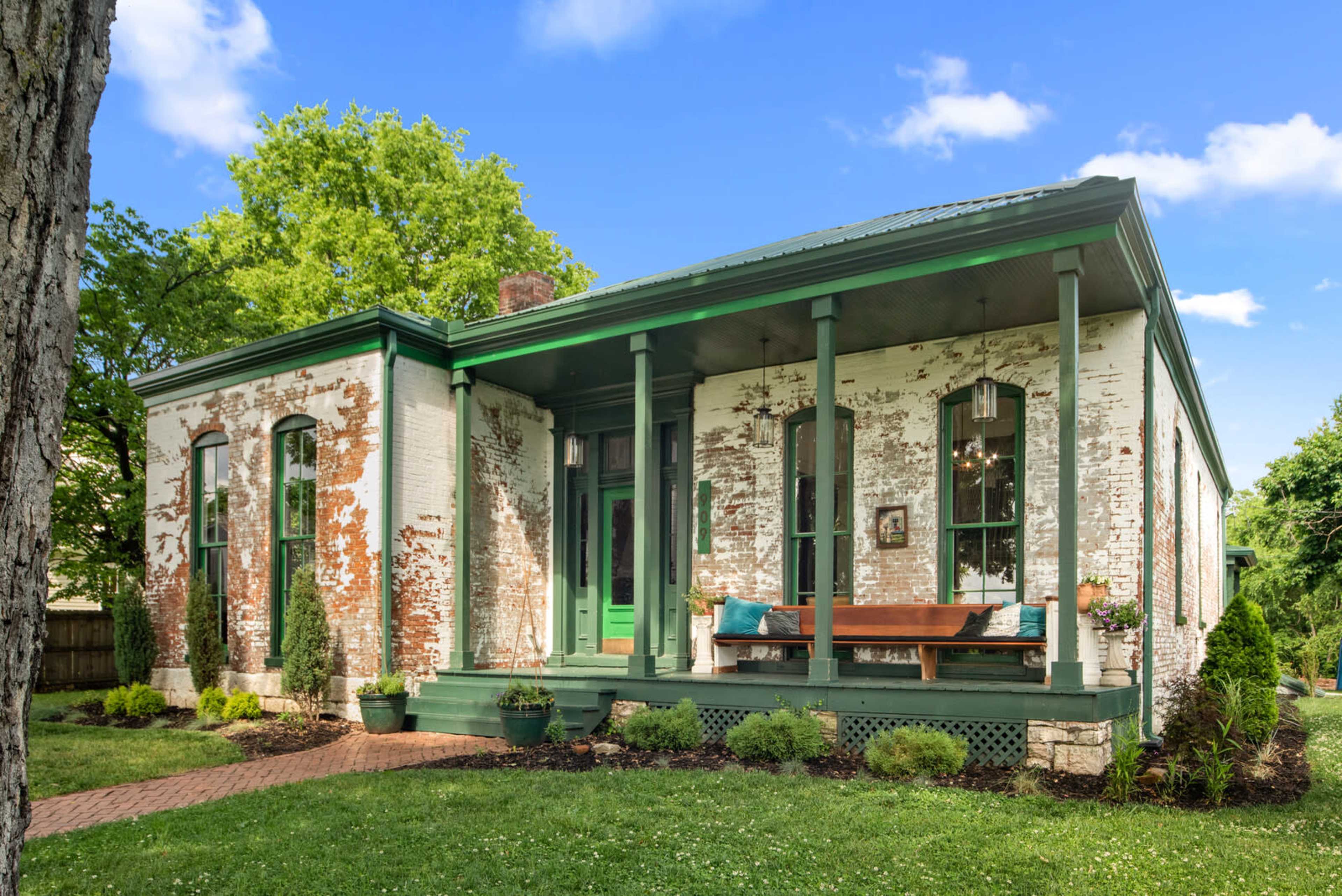 A brick house with green accents, featuring large windows and a front porch with seating, surrounded by lush greenery.