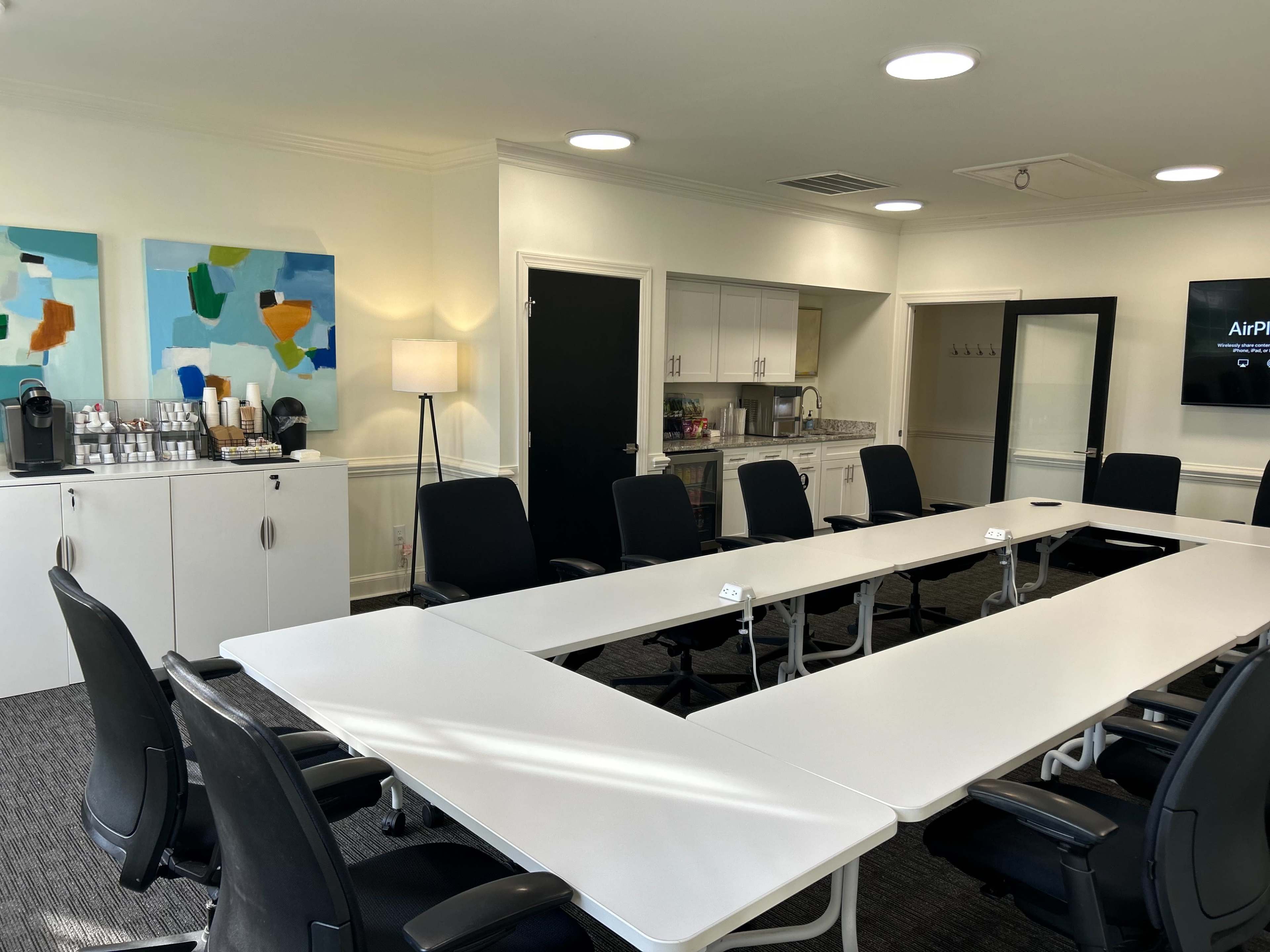 The image shows a modern conference room with a long white table surrounded by black chairs, featuring coffee supplies and artwork on the walls.