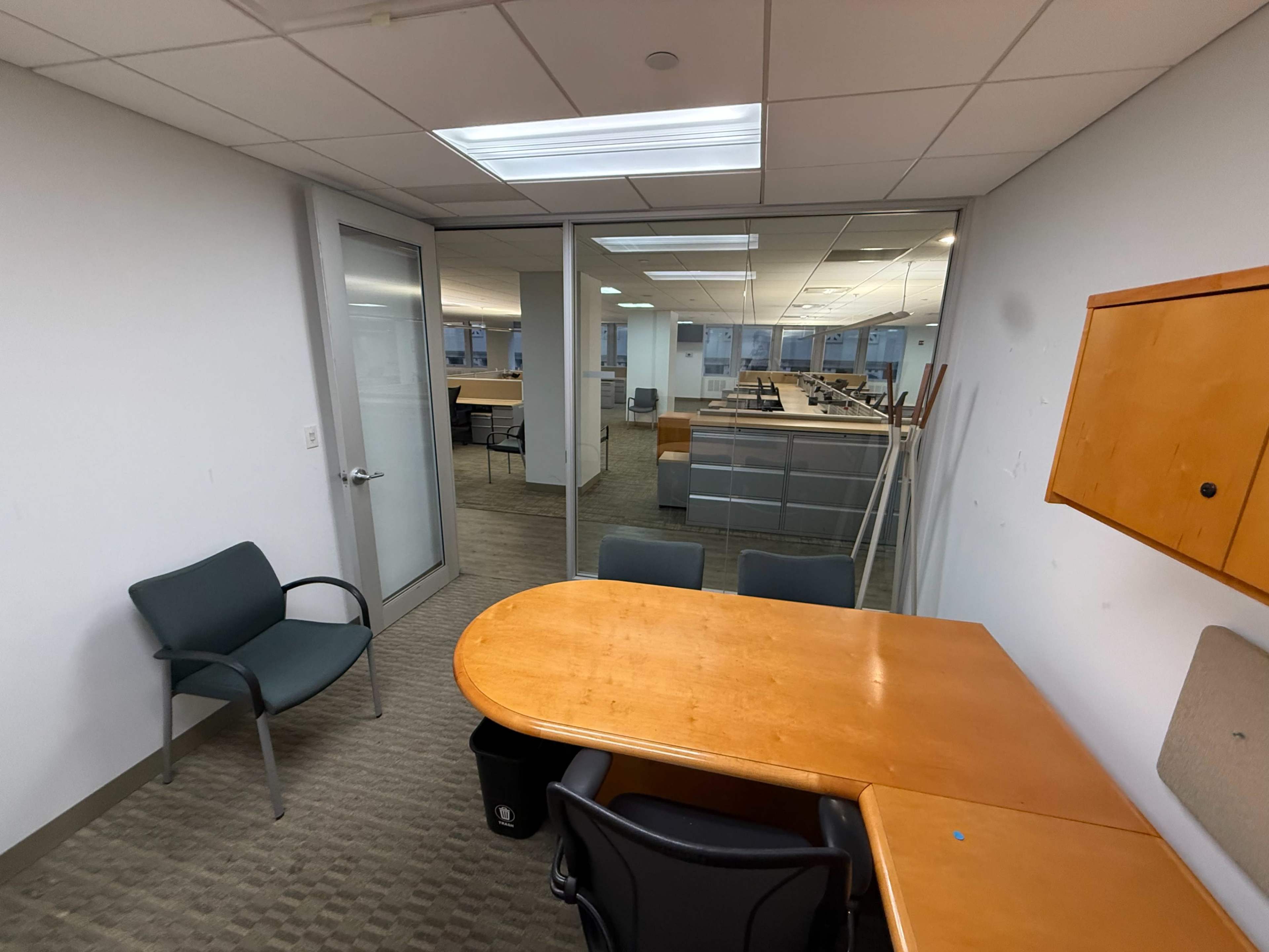The image shows a small office space with a round wooden table and two chairs, partially separated by a glass wall from a larger, empty workspace filled with desks.