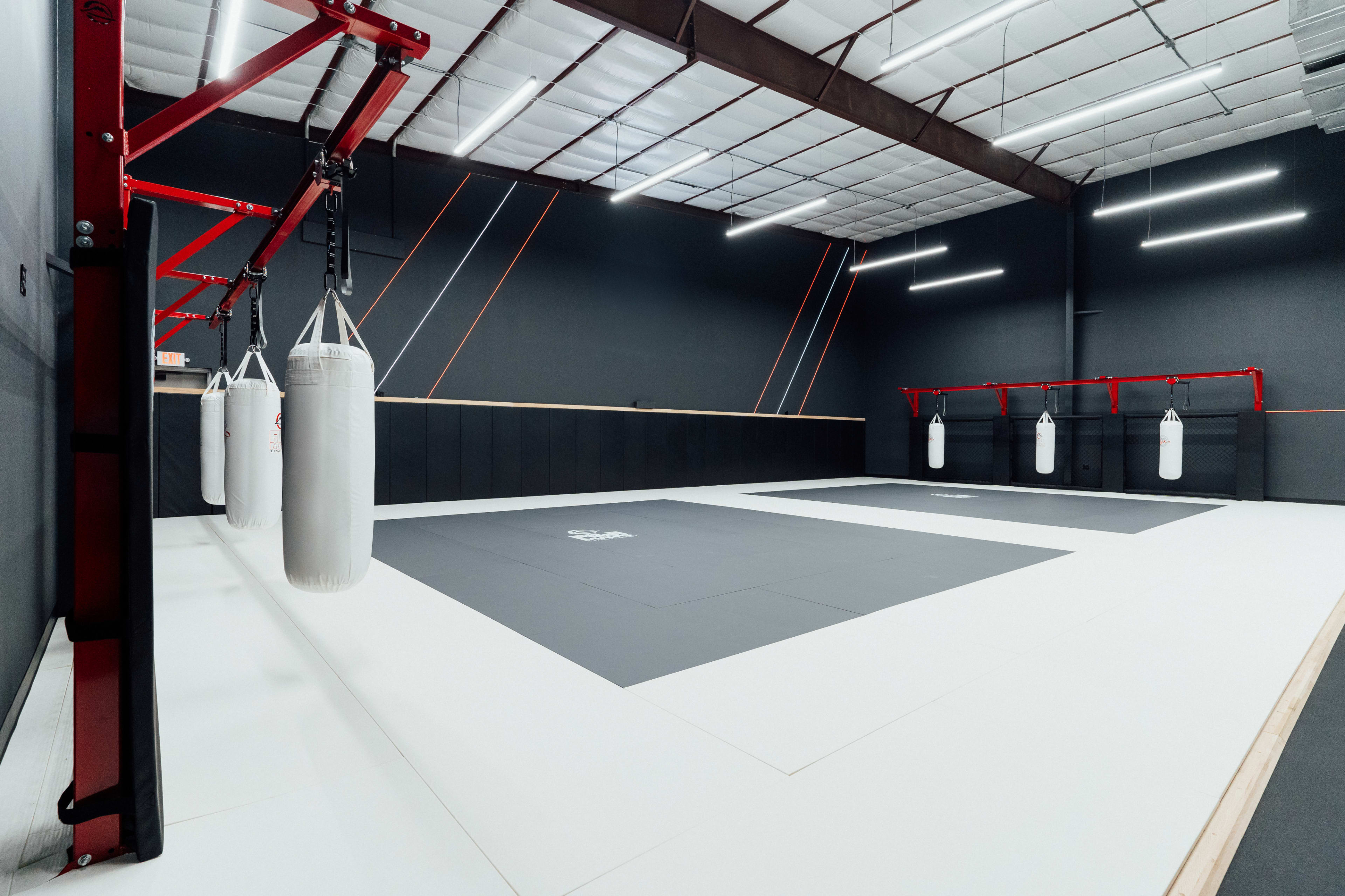 A martial arts training facility with white and gray mats, four hanging punching bags, and a spacious, uncluttered layout.