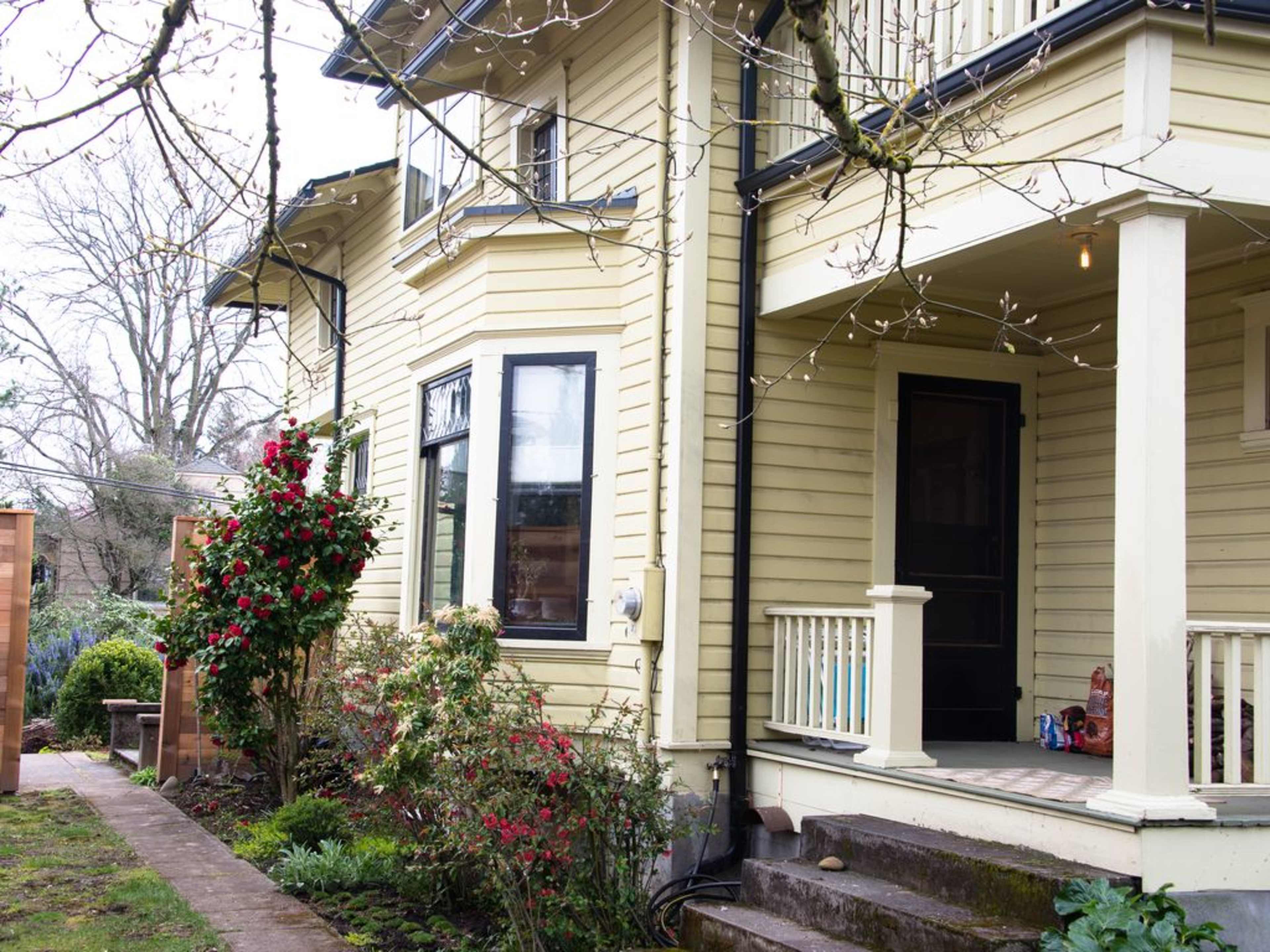 A yellow house with a porch features a garden with flowers and a pathway leading to its entrance.